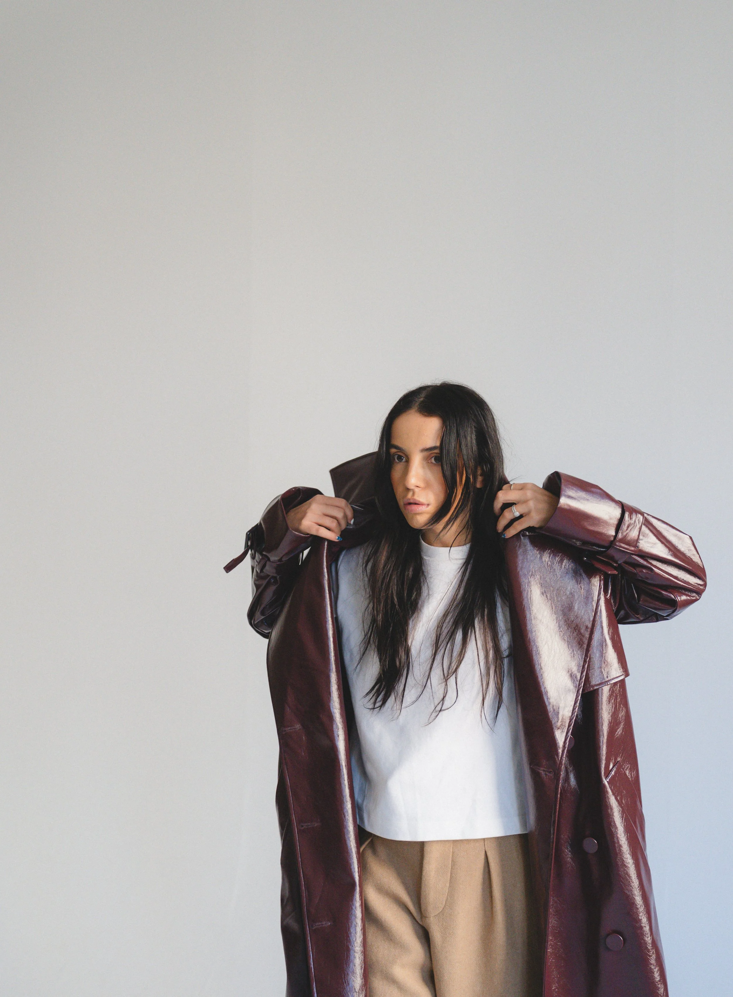 A woman with long dark hair looking at the camera, wearing a white shirt and beige pants, with a large maroon satin trench coat draped over her shoulders, standing against a plain light gray background.