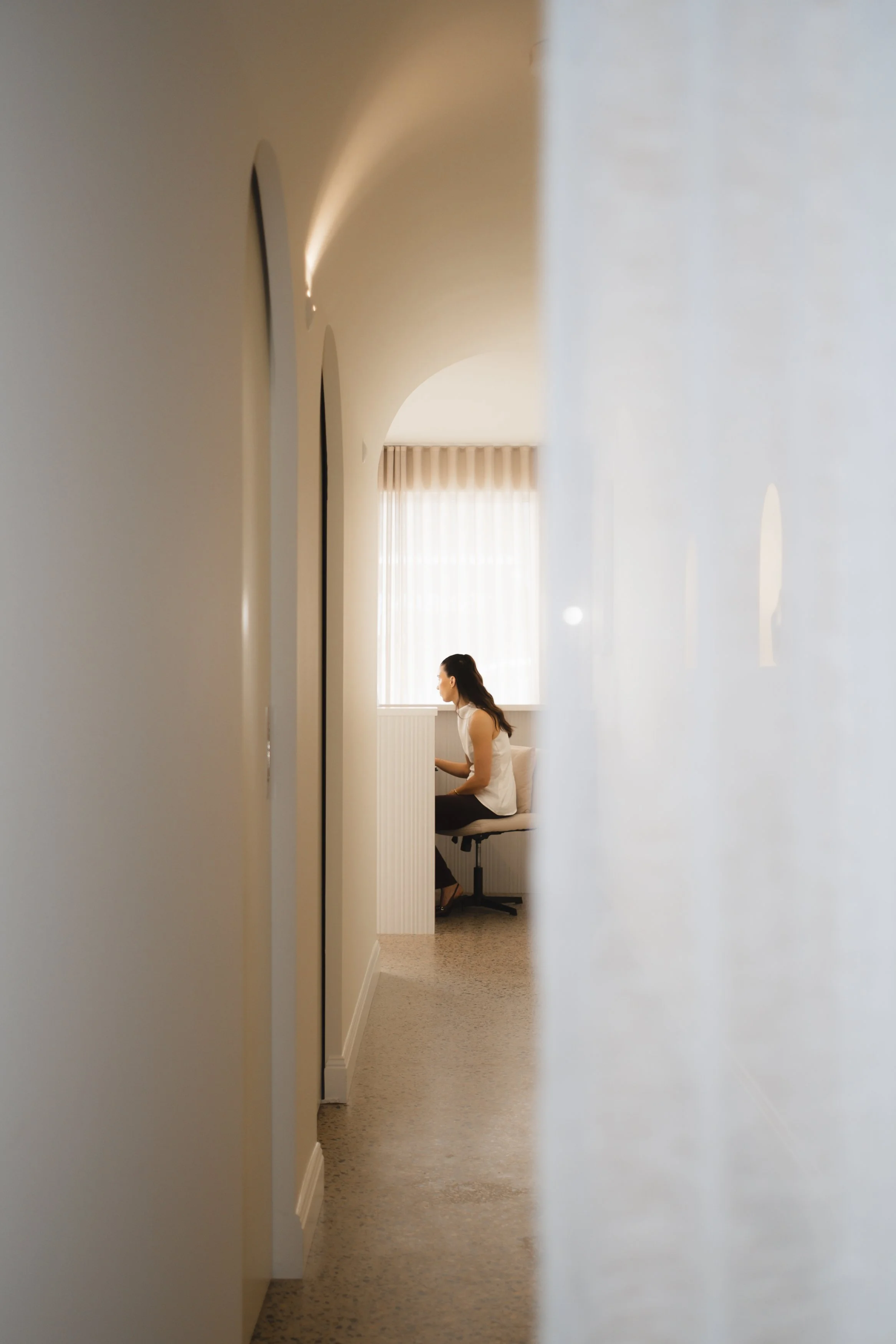 A woman sitting at a desk near a window with curtains, framed by an indoor wall and a door frame.