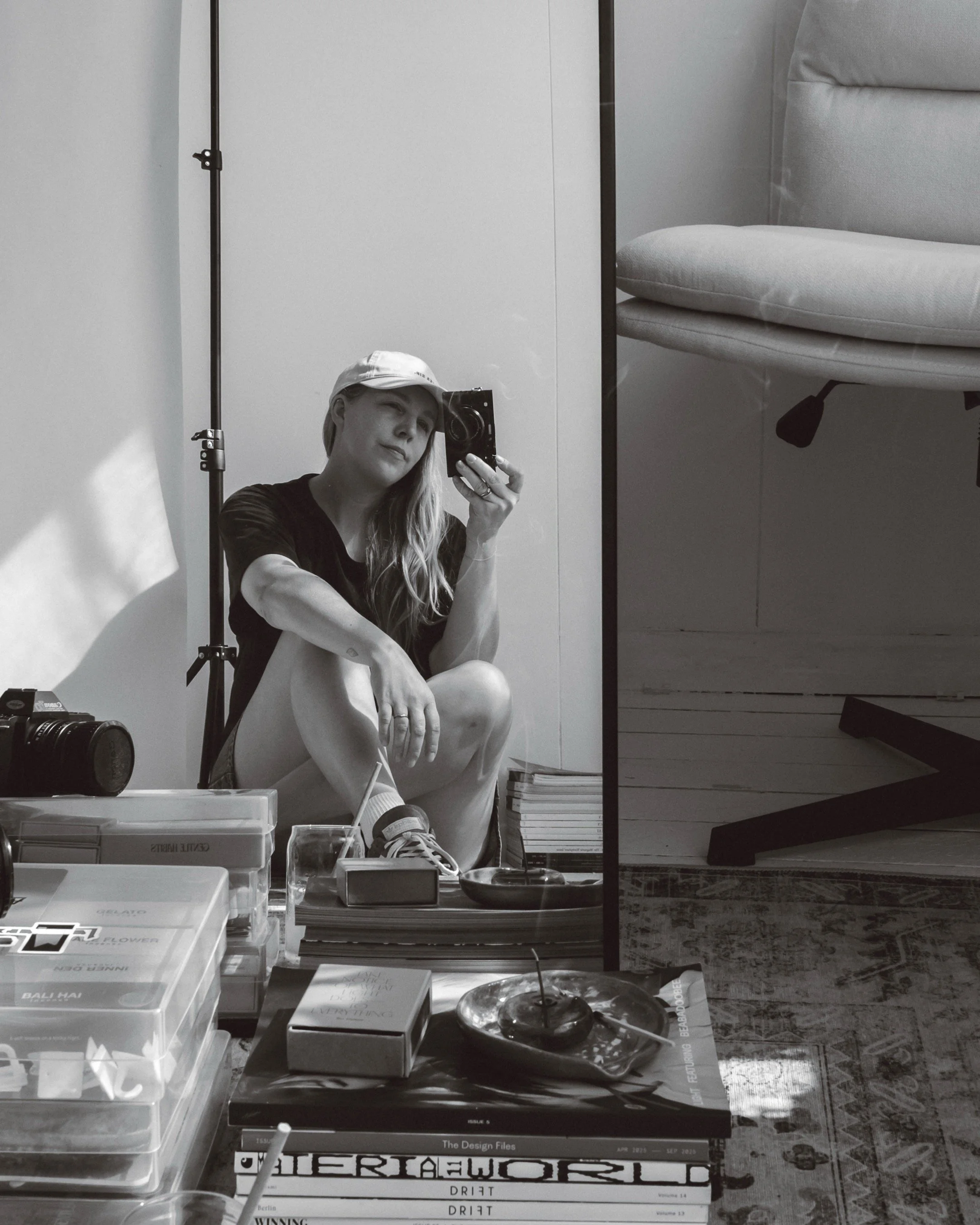 A woman taking a mirror selfie in a room with a camera on the table, books, a donut with a candle, and various items on a table, sitting on the floor with a casual pose.