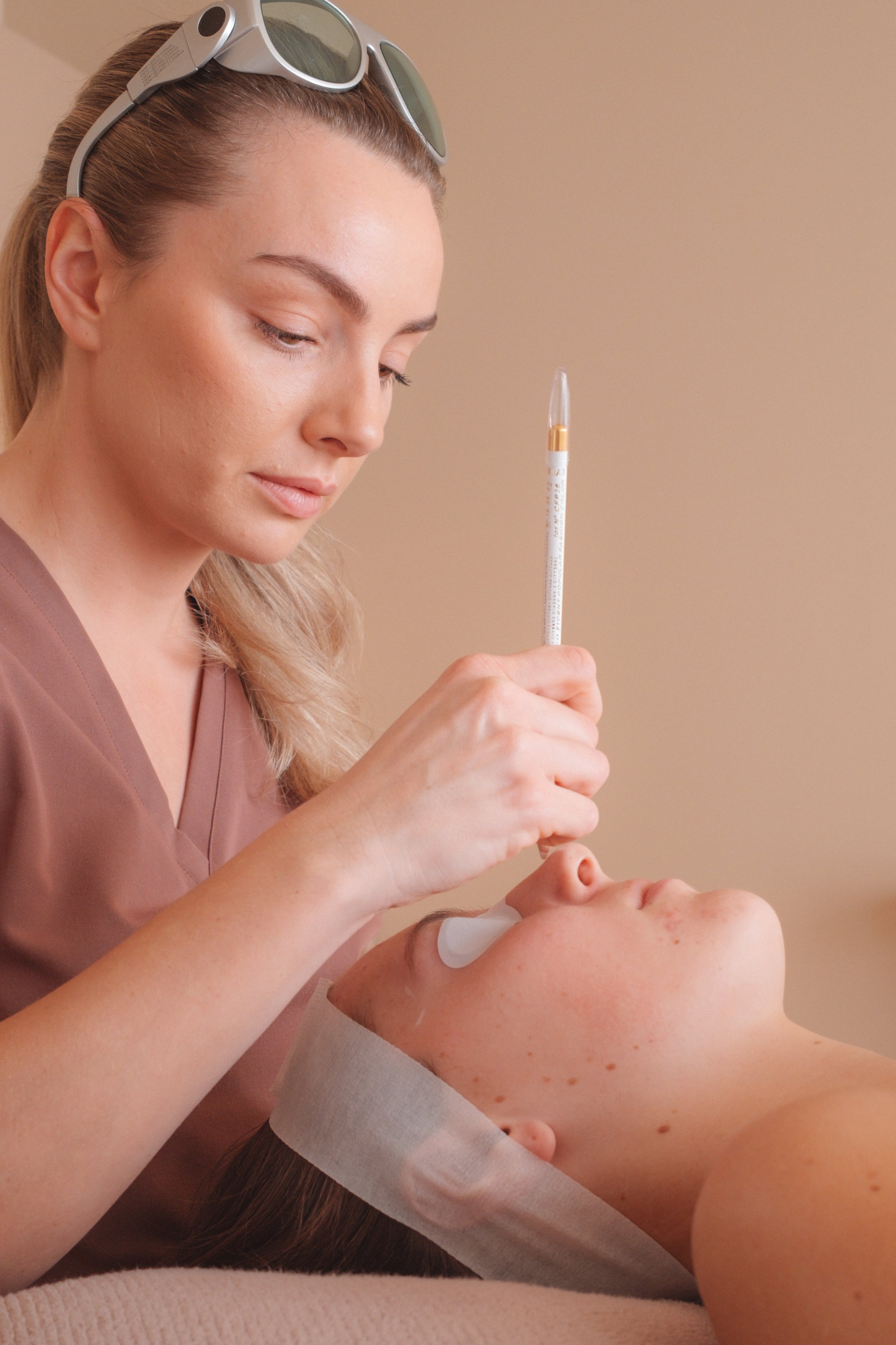 A cosmetologist performs a facial treatment on a client lying down with a facial mask on. The cosmetologist is holding a syringe and wearing glasses resting on her head. The client has facial redness and acne.