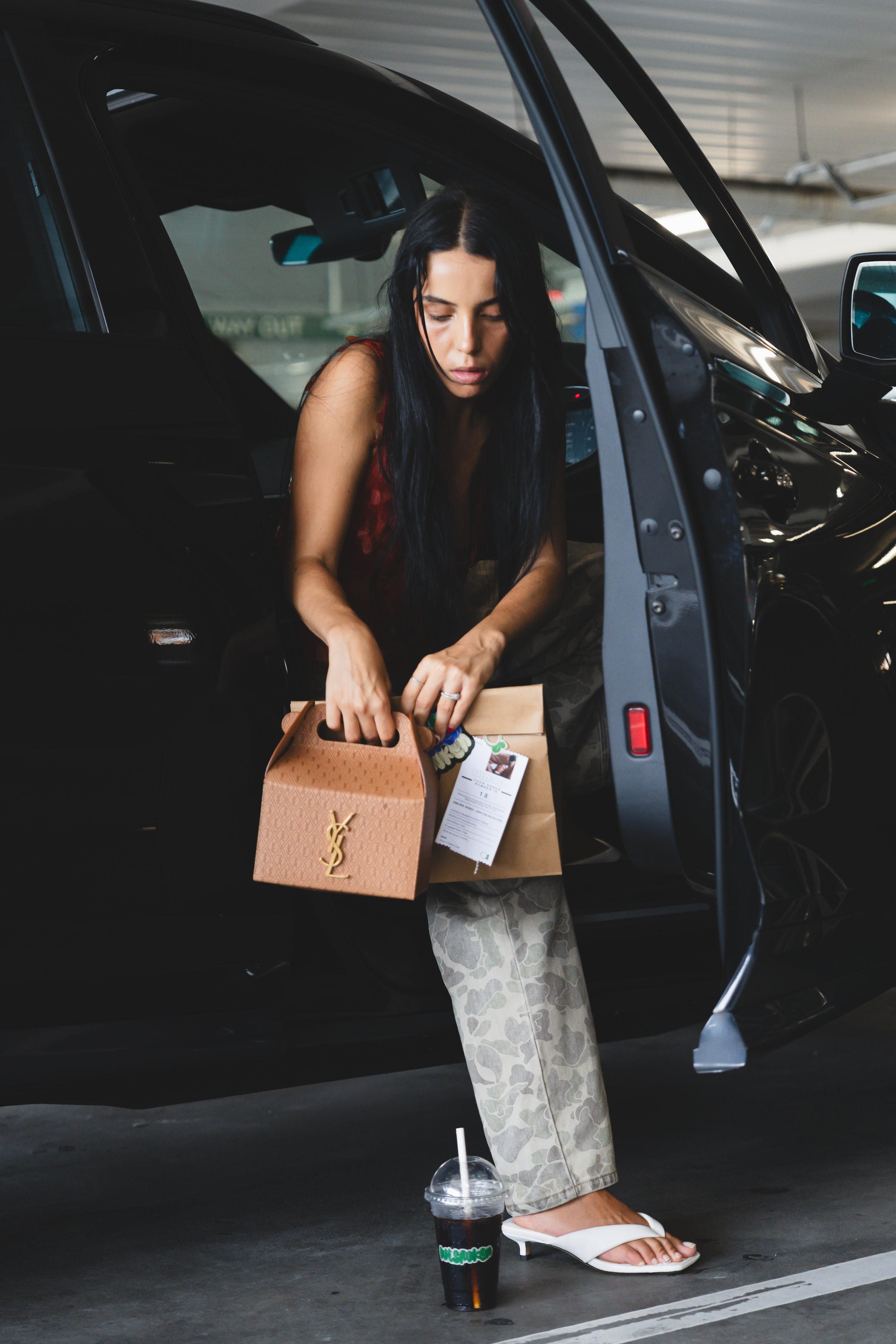 A woman with long black hair leaning into a black car, holding a shopping bag and a paper bag inside the vehicle. She is wearing patterned pants and white high-heeled sandals. On the ground outside the car, there is a drink in a plastic cup with a st