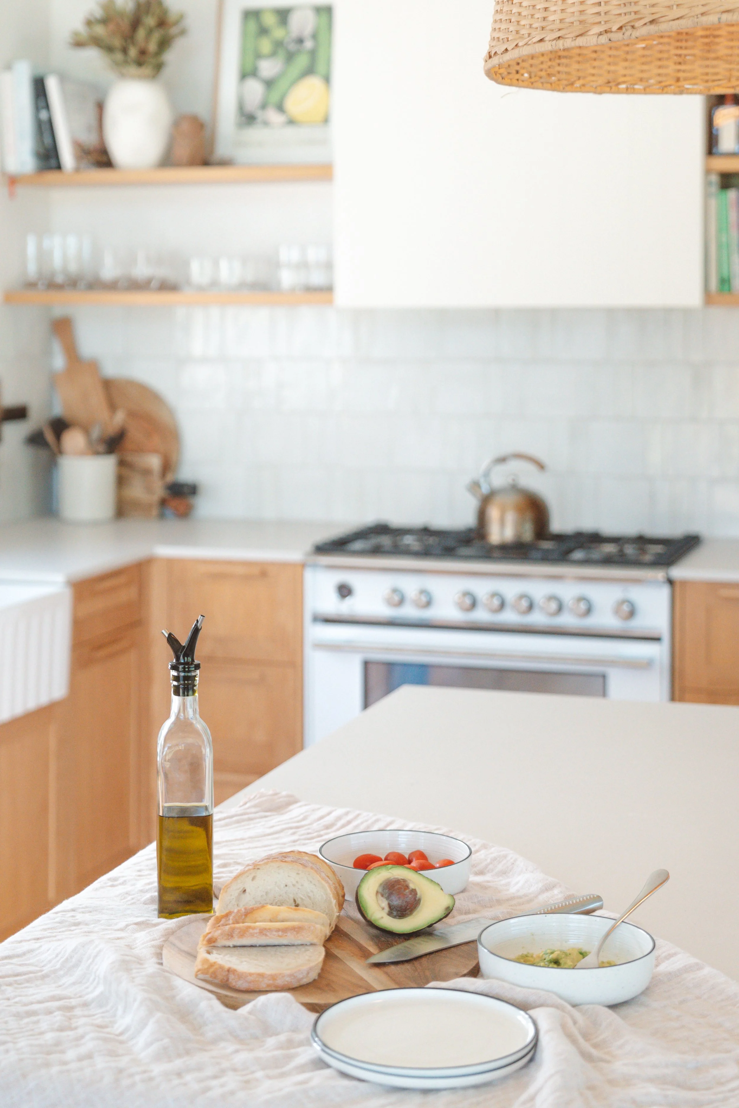 A breakfast table with sliced bread, avocado, cherry tomatoes, a bowl of soup, and a bottle of olive oil with a pour spout in a bright kitchen.