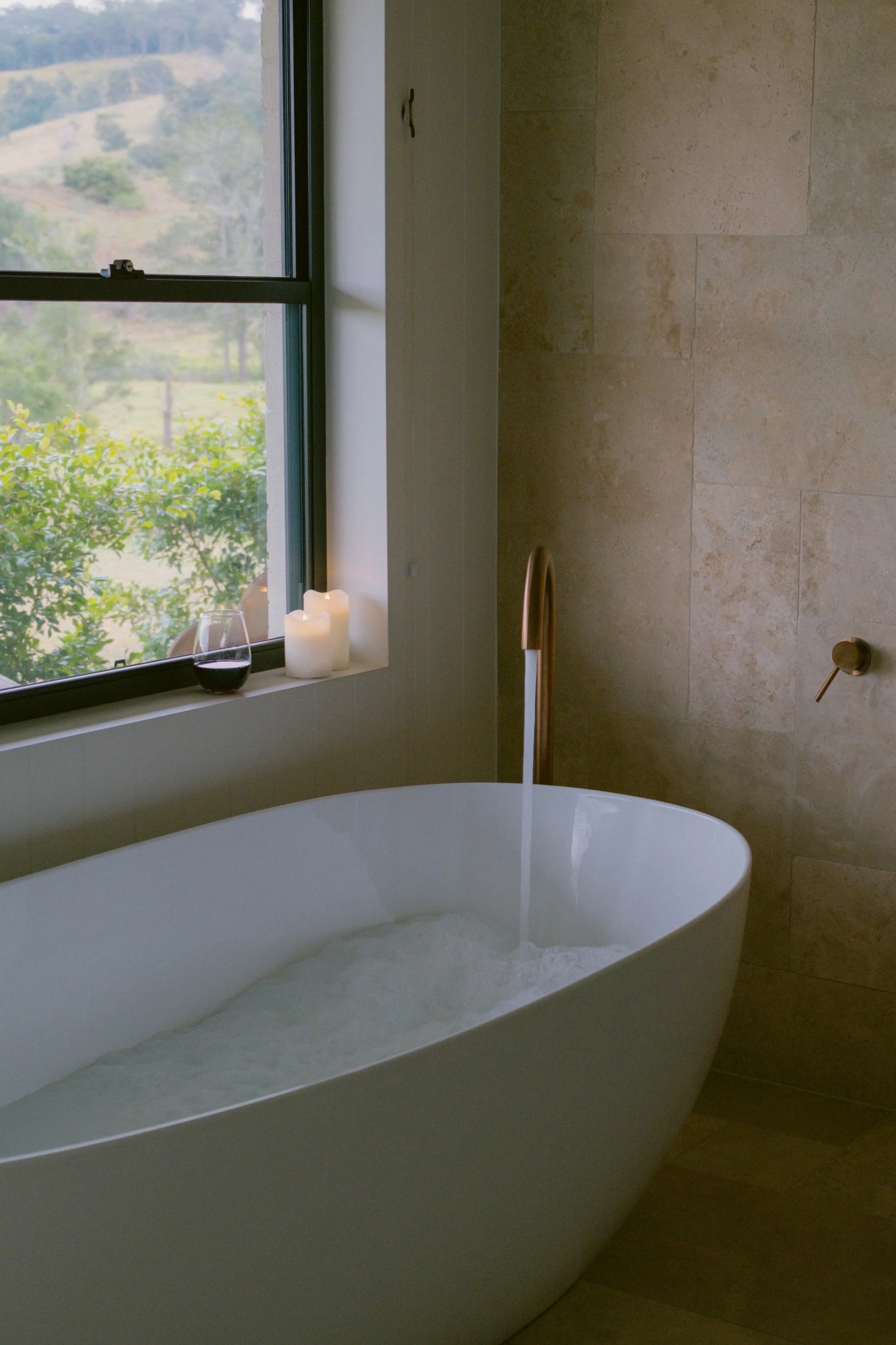A bathroom with a freestanding bathtub filled with water, a window with a view of greenery and hills outside, lit candles and a glass of red wine on the windowsill, and a gold-colored faucet with water flowing into the tub.