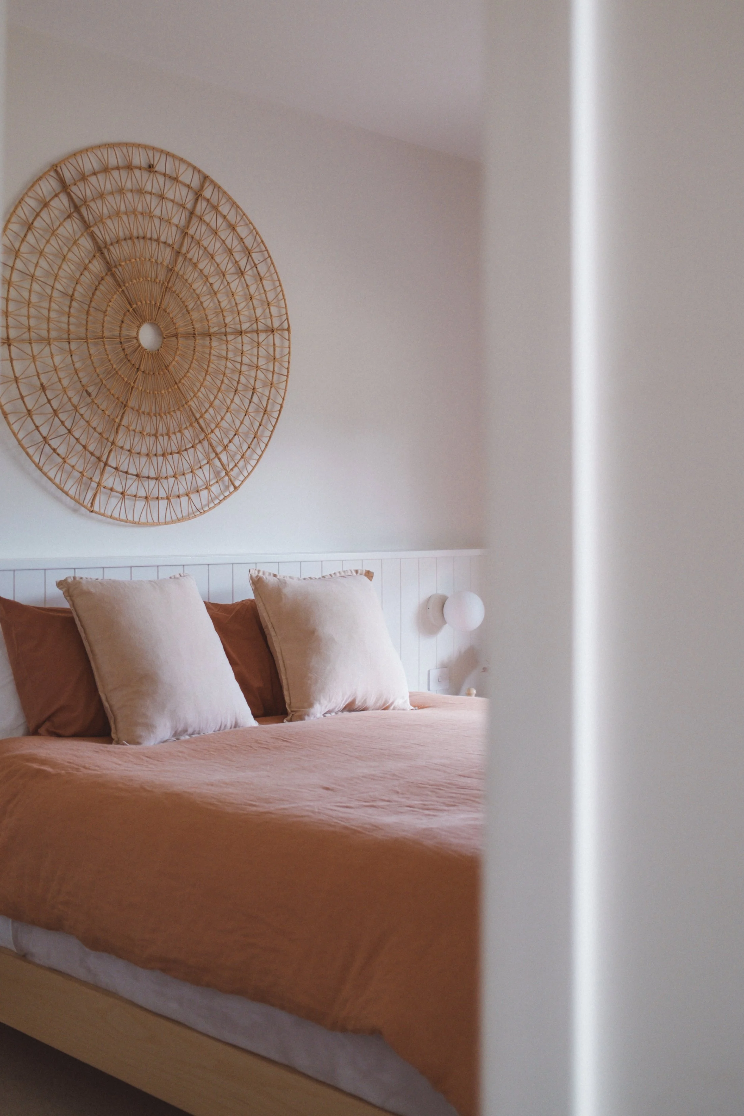 A bedroom with a bed covered in a peach-colored bedspread and four beige pillows. A white headboard is visible. Behind the bed, a round, woven wall decoration is on the wall, and a small wall-mounted lamp is also visible.