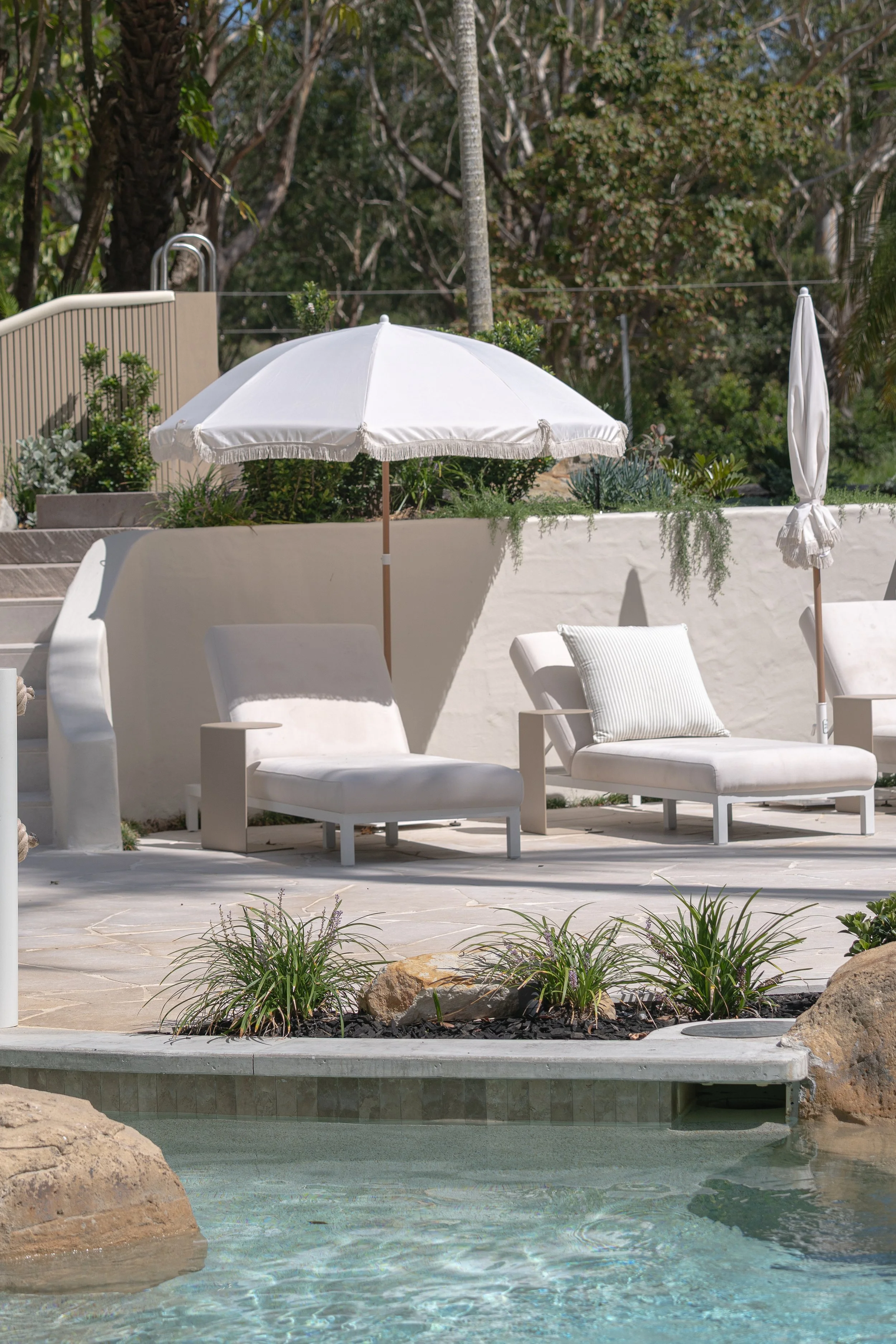 Photo of a backyard pool area with white lounge chairs, patio umbrellas, lush greenery, and a small waterfall feature with rocks and plants.