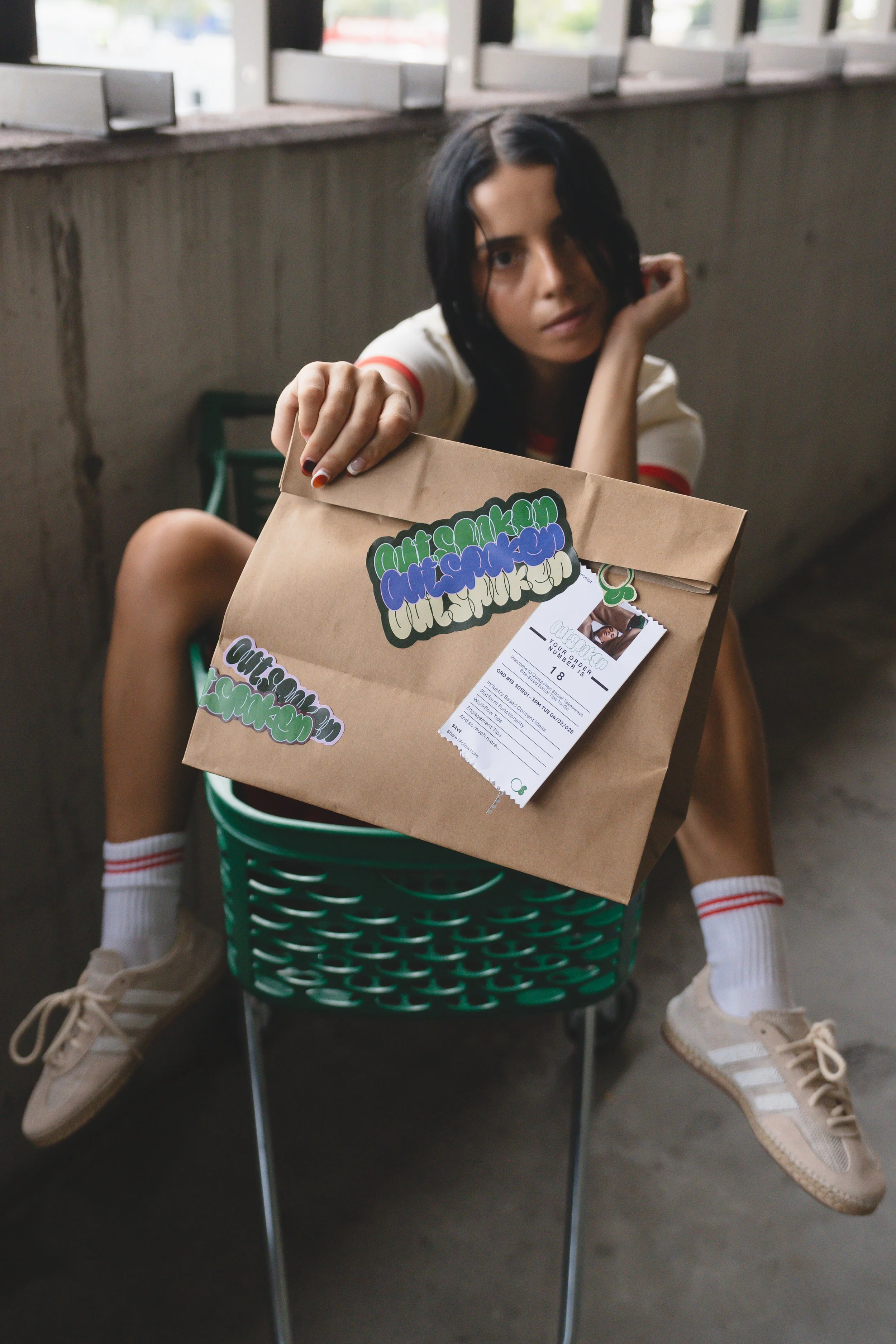 A person with black hair and light skin sitting in a green shopping cart, holding a brown paper bag with colorful graffiti-style stickers, wearing beige sneakers and white socks with red stripes, inside a building with large windows.