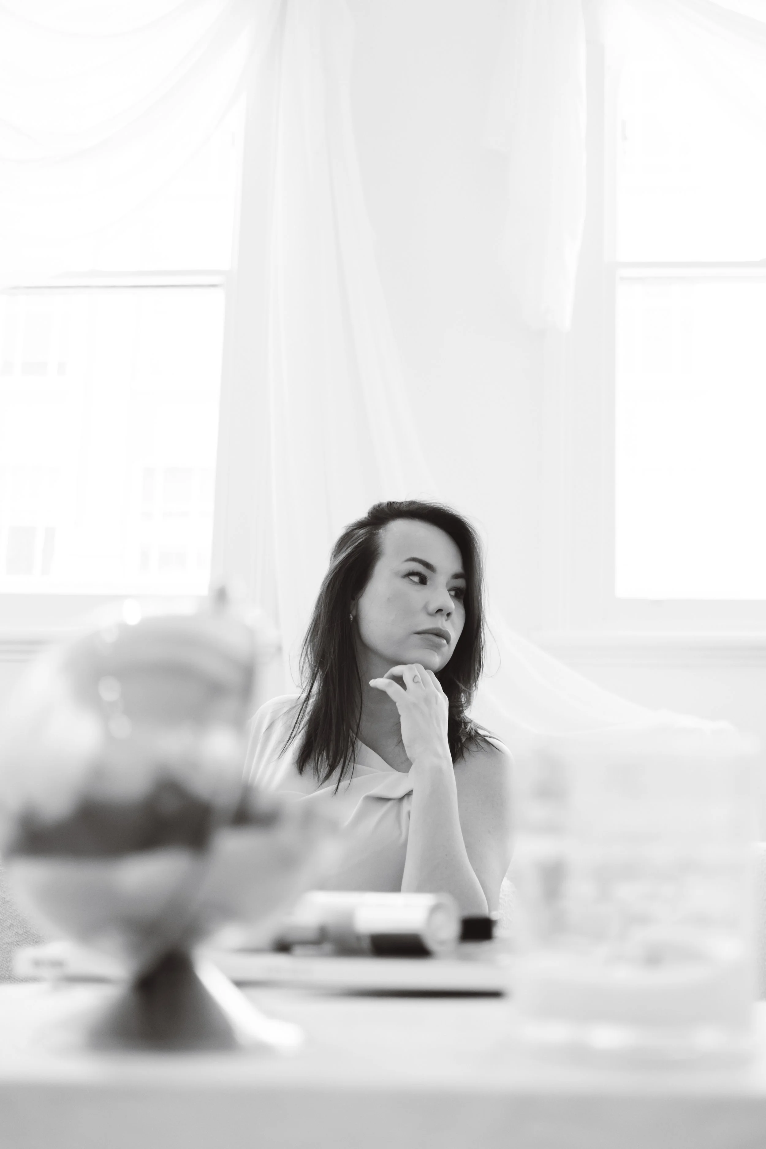 Black and white photo of a woman with long hair sitting at a table, with her chin resting on her hand, in a bright room with large windows and curtains.
