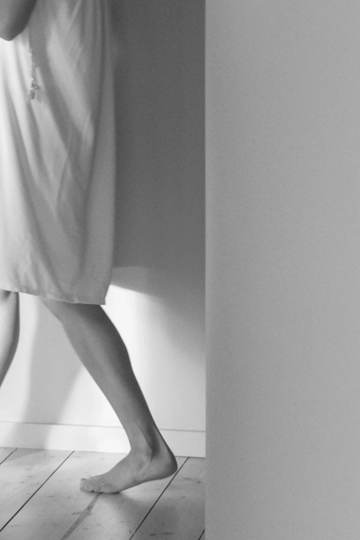 A person standing barefoot on a tiled floor, wearing a loose dress, near a wall and curtain in a minimalistic room.