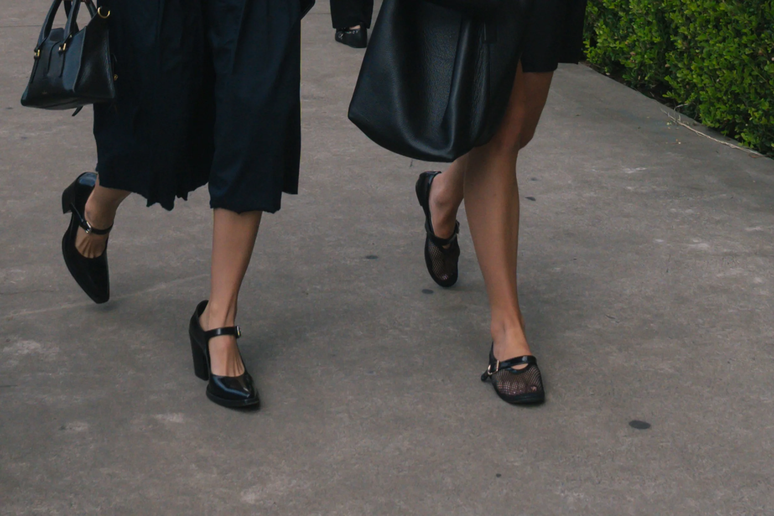Close-up of women's legs and feet walking on sidewalk, wearing black shoes and sheer stockings, with black handbags.