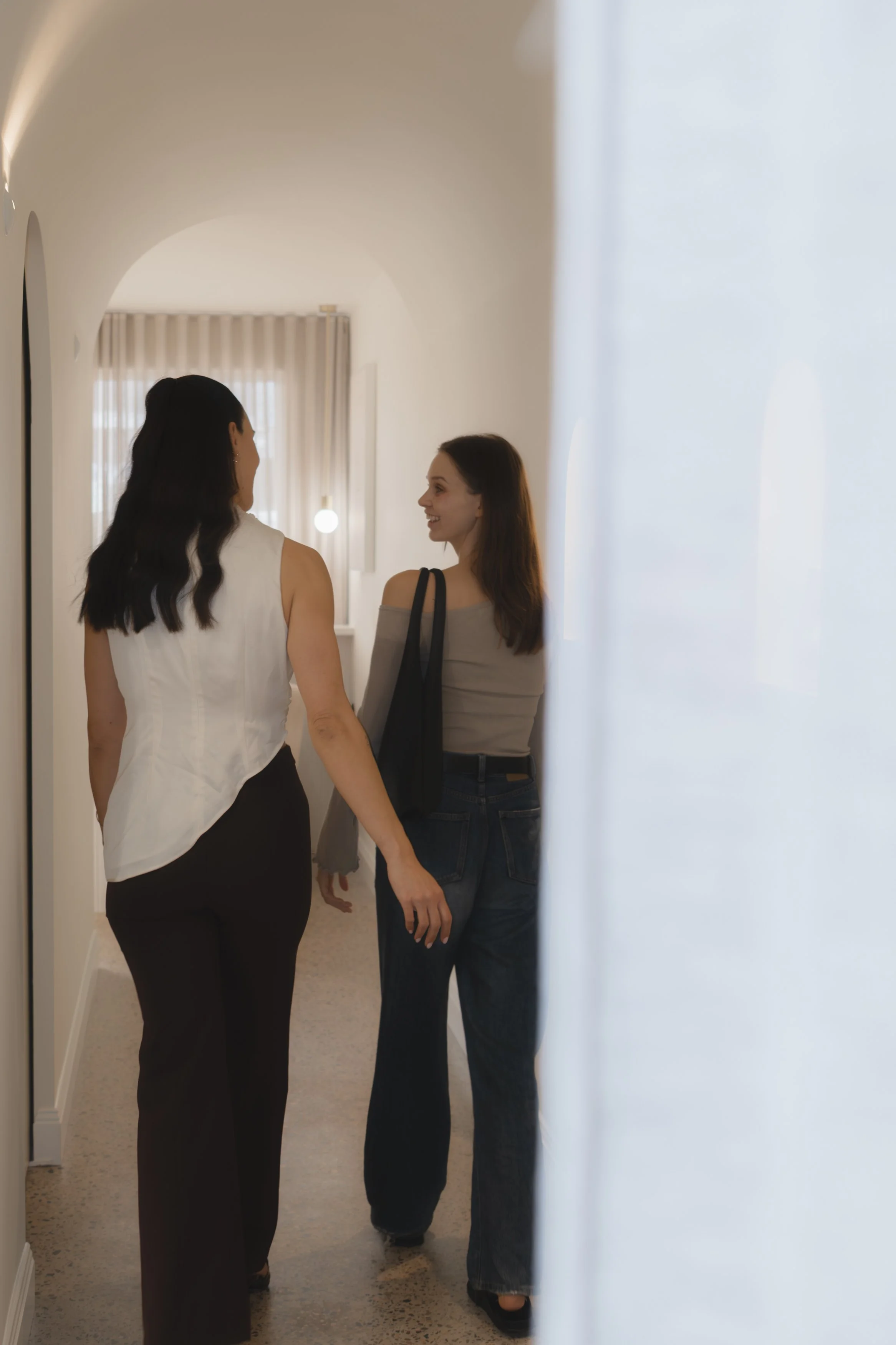 Two women are walking down a hallway and talking to each other. One has dark hair and is wearing a sleeveless white top and dark pants, while the other has brown hair and is dressed in a grey off-the-shoulder top and jeans.