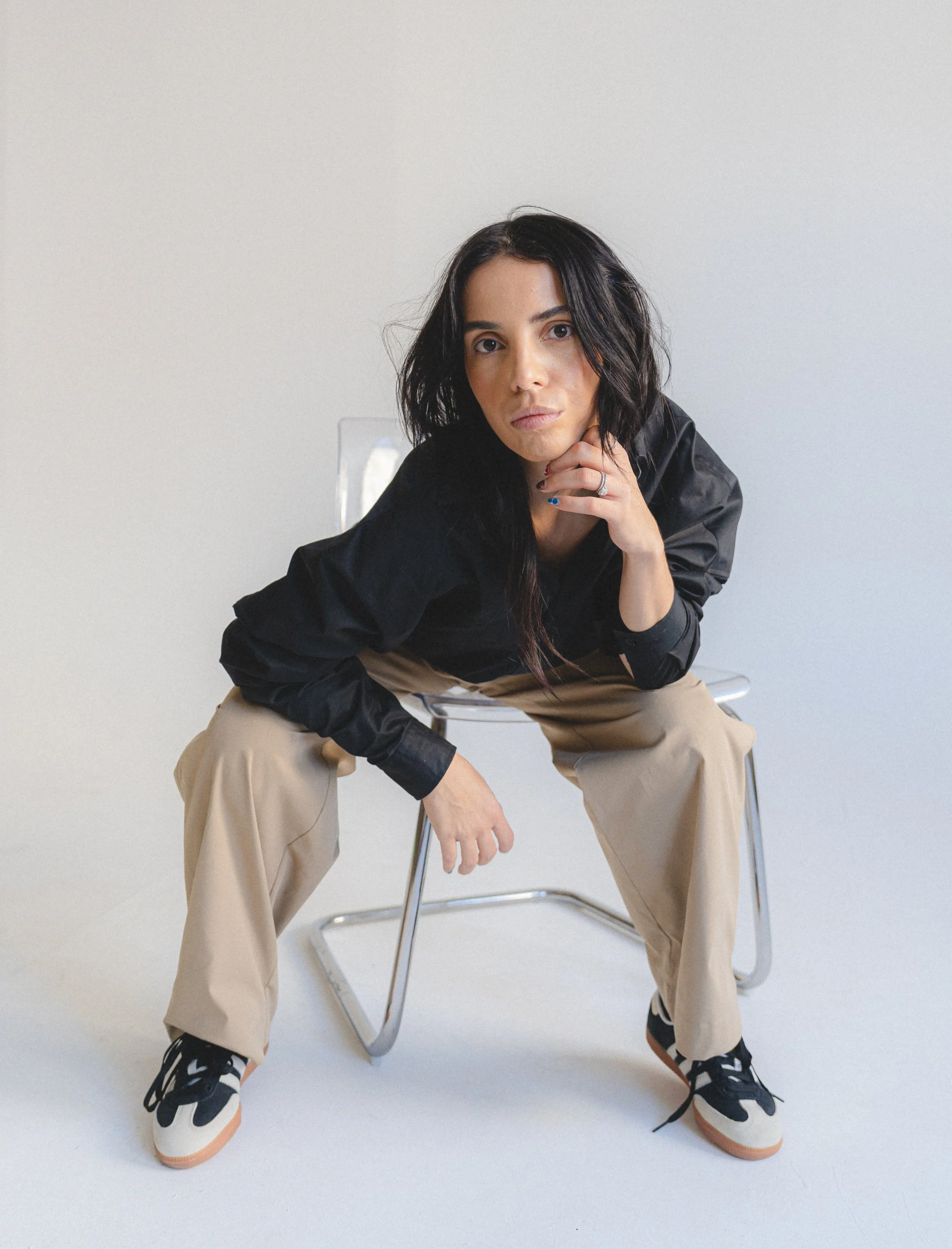 A woman with dark hair, wearing a black jacket, khaki pants, and sneakers, sitting on a metal chair against a plain white background, with her chin resting on her hand.