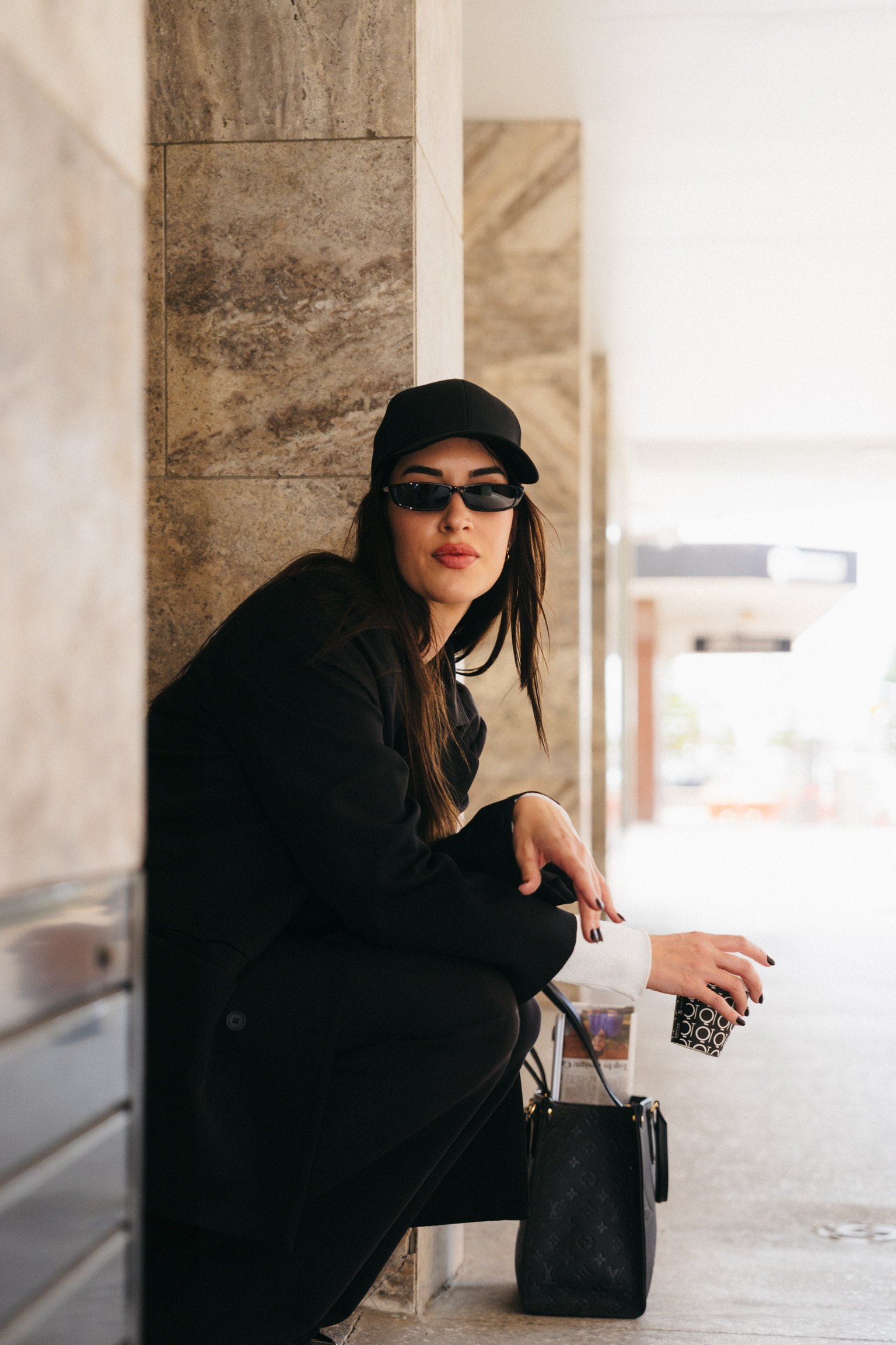 A woman wearing a black cap, black sunglasses, and a black jacket, sitting against a stone wall in a well-lit indoor space. She has long dark hair, neutral lipstick, and is holding a drink with a black and white pattern. There is a black Louis Vuitto