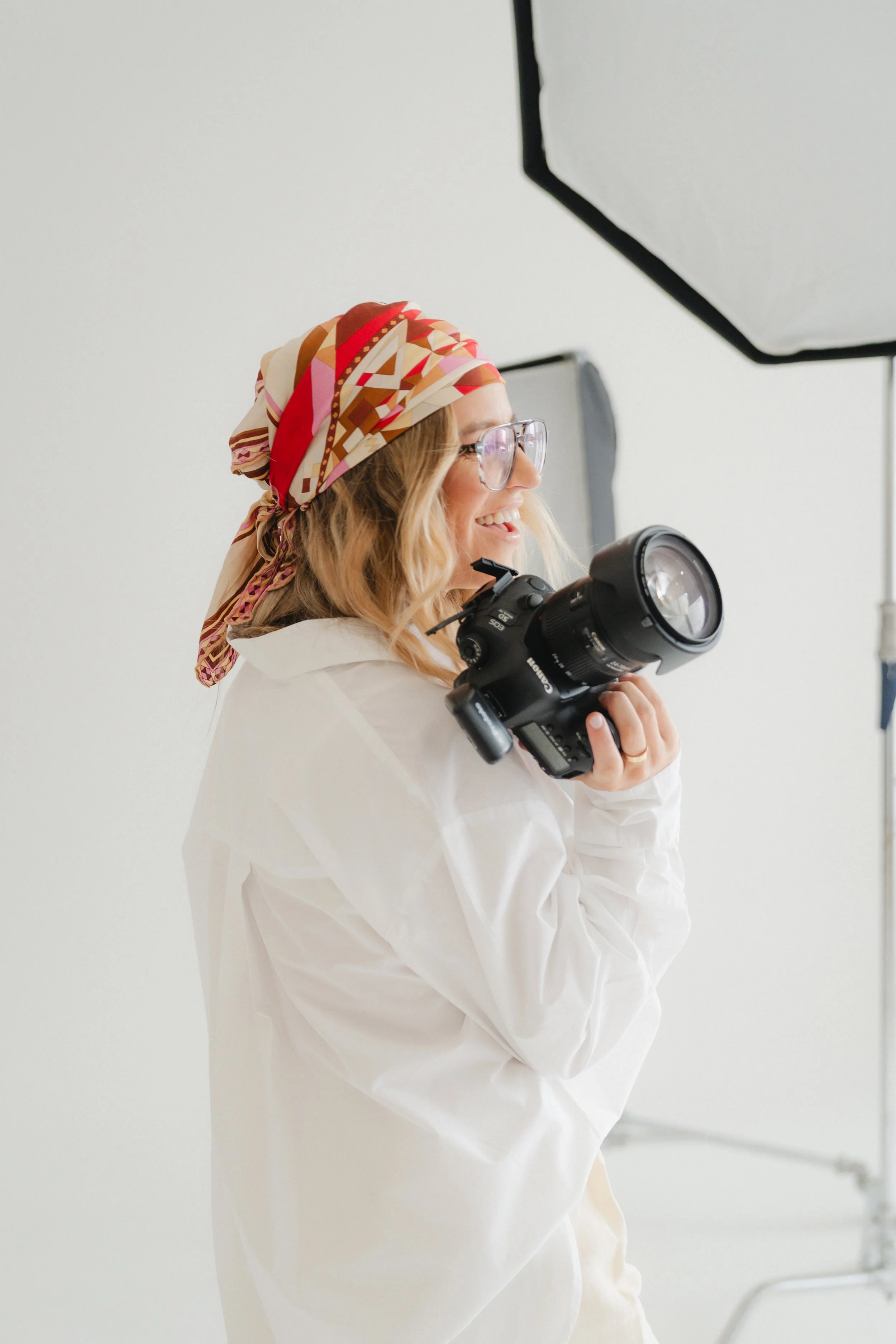 A woman with blonde hair, glasses, and a colorful headscarf smiling and holding a professional camera while standing against a plain white background.