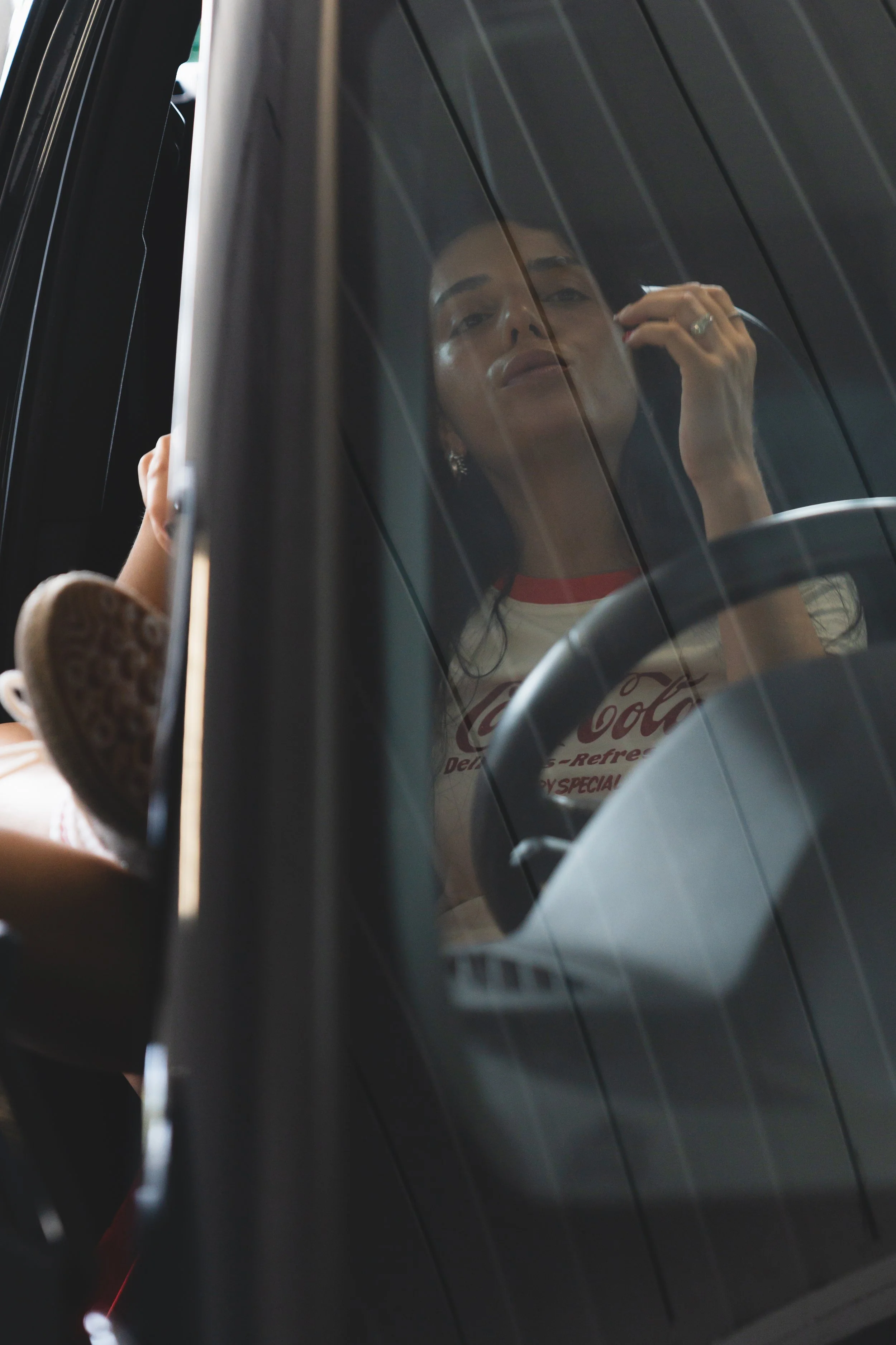 A woman with dark hair and earrings is looking at herself in a reflection on a car window while touching her face, with a hand with rings visible. She is wearing a white shirt with a logo and red collar.