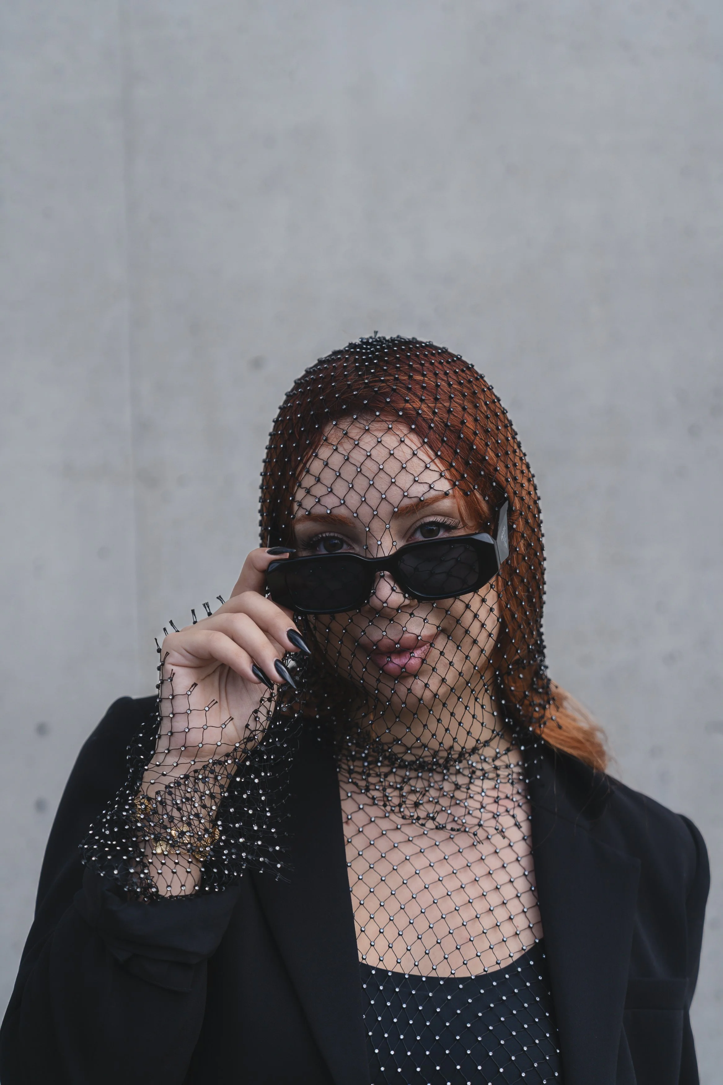 A woman with reddish hair, wearing a fishnet top and blazer, holds black sunglasses with one hand, partially covering her face, against a plain light-colored background.