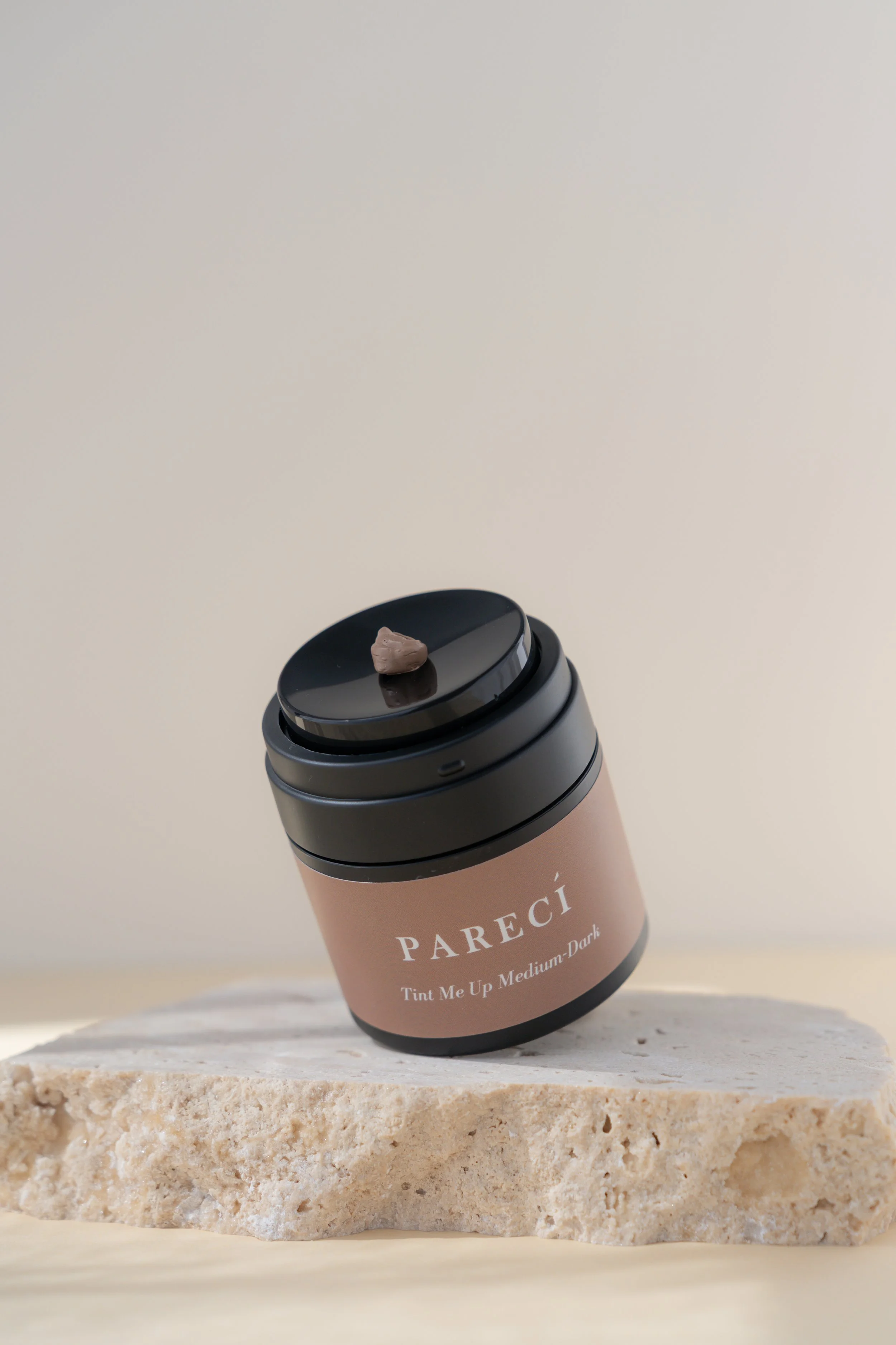 A jar of PARECI Tint Me Up Medium-Dark makeup cream with a small amount of product on the black lid, placed on a light-colored, textured stone surface against a plain cream background.