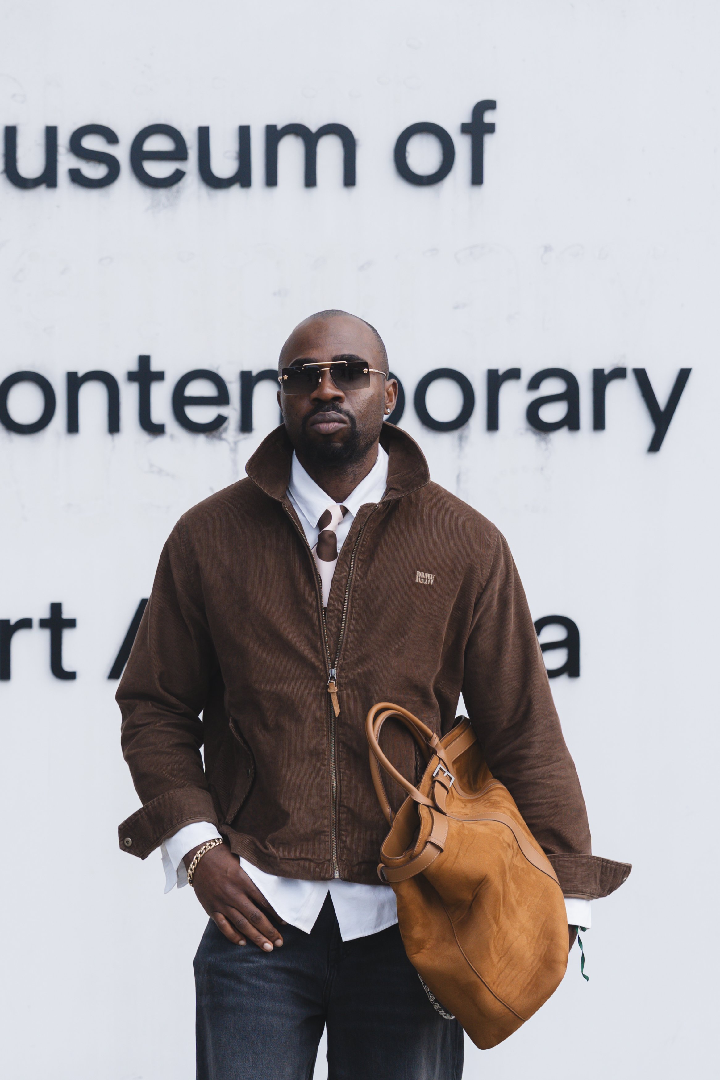 A man wearing sunglasses, a brown jacket, and a white shirt, holding a tan bag, stands in front of a white wall with black text that reads 'museum of' and 'ponterary'.
