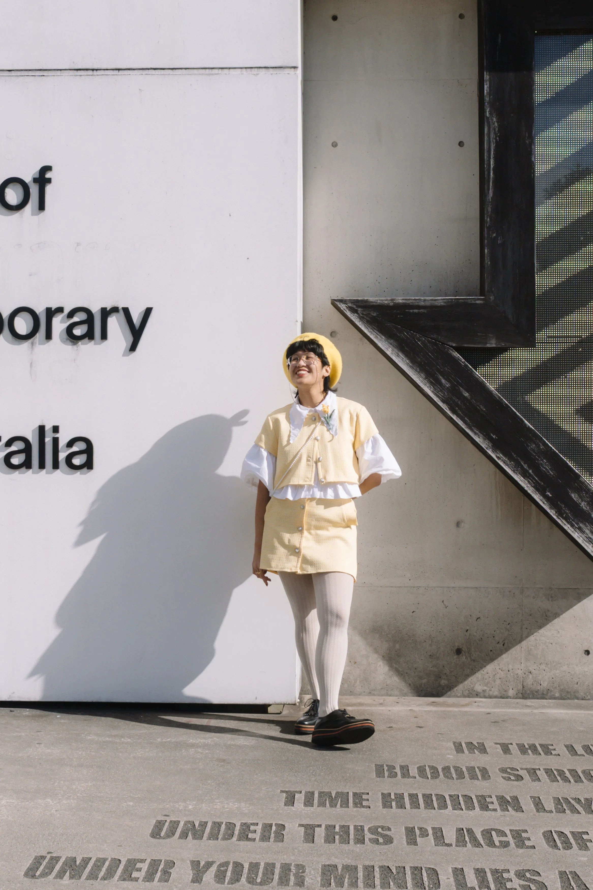 A woman wearing a yellow hat and beige outfit standing in front of a building with text on the ground, smiling and looking upward.