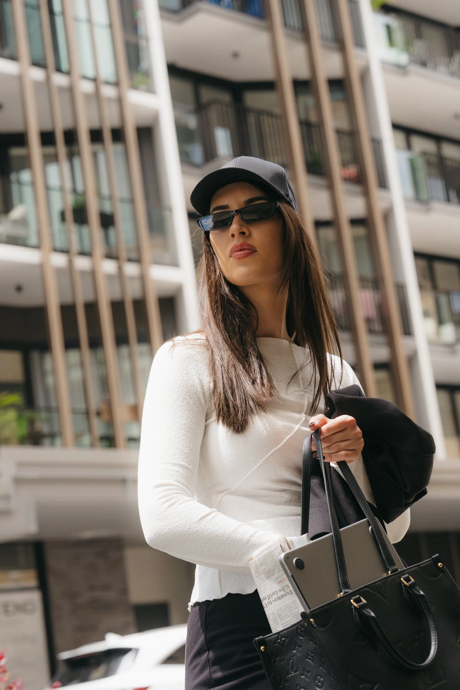 A woman with long brown hair, wearing a black baseball cap, black sunglasses, a white sweater, and black pants, stands outdoors holding a large designer black handbag and a newspaper. Behind her is a multi-story building with balconies and wooden ver