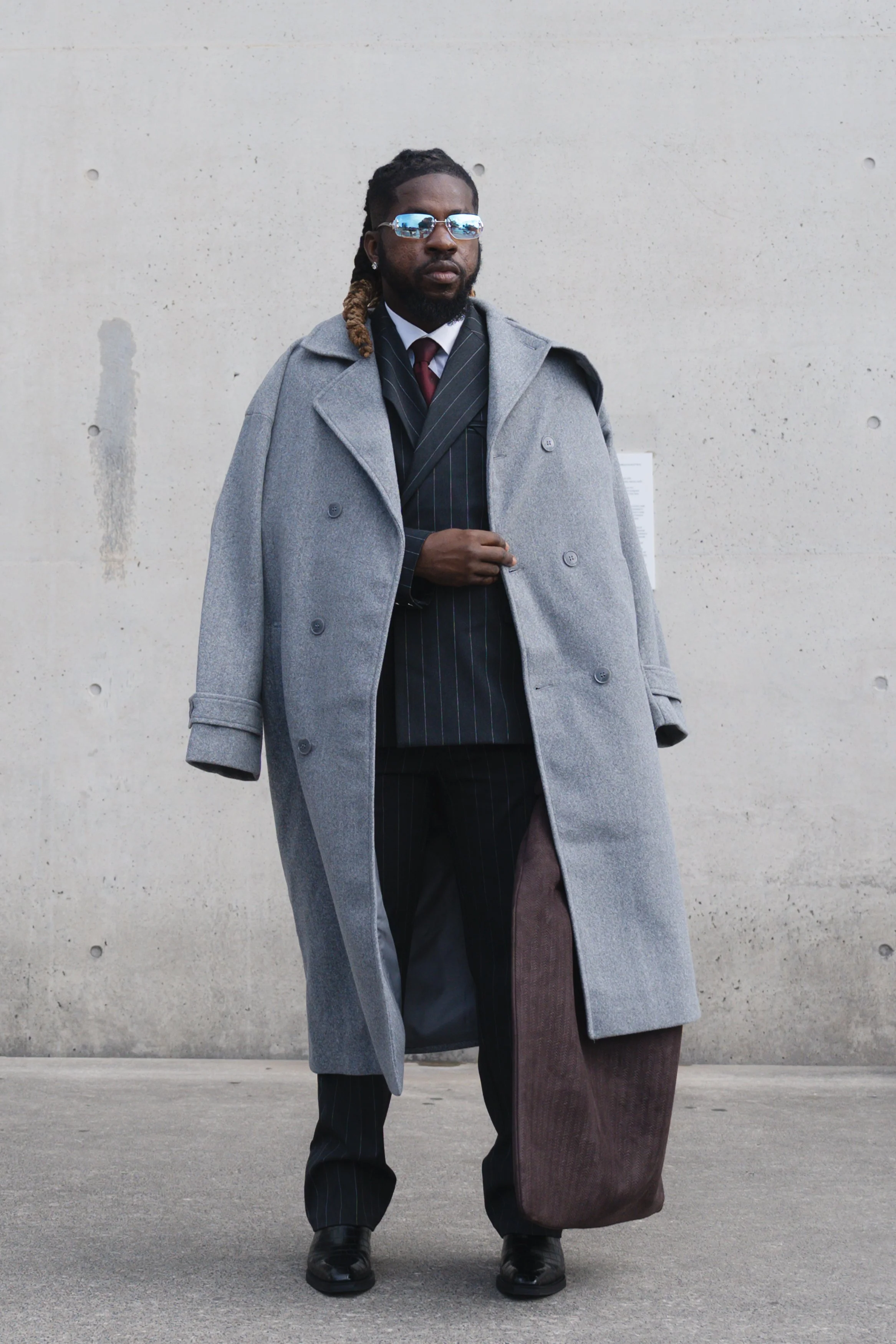 A man wearing a long grey coat, sunglasses, a suit, and holding a bag, standing against a beige wall.