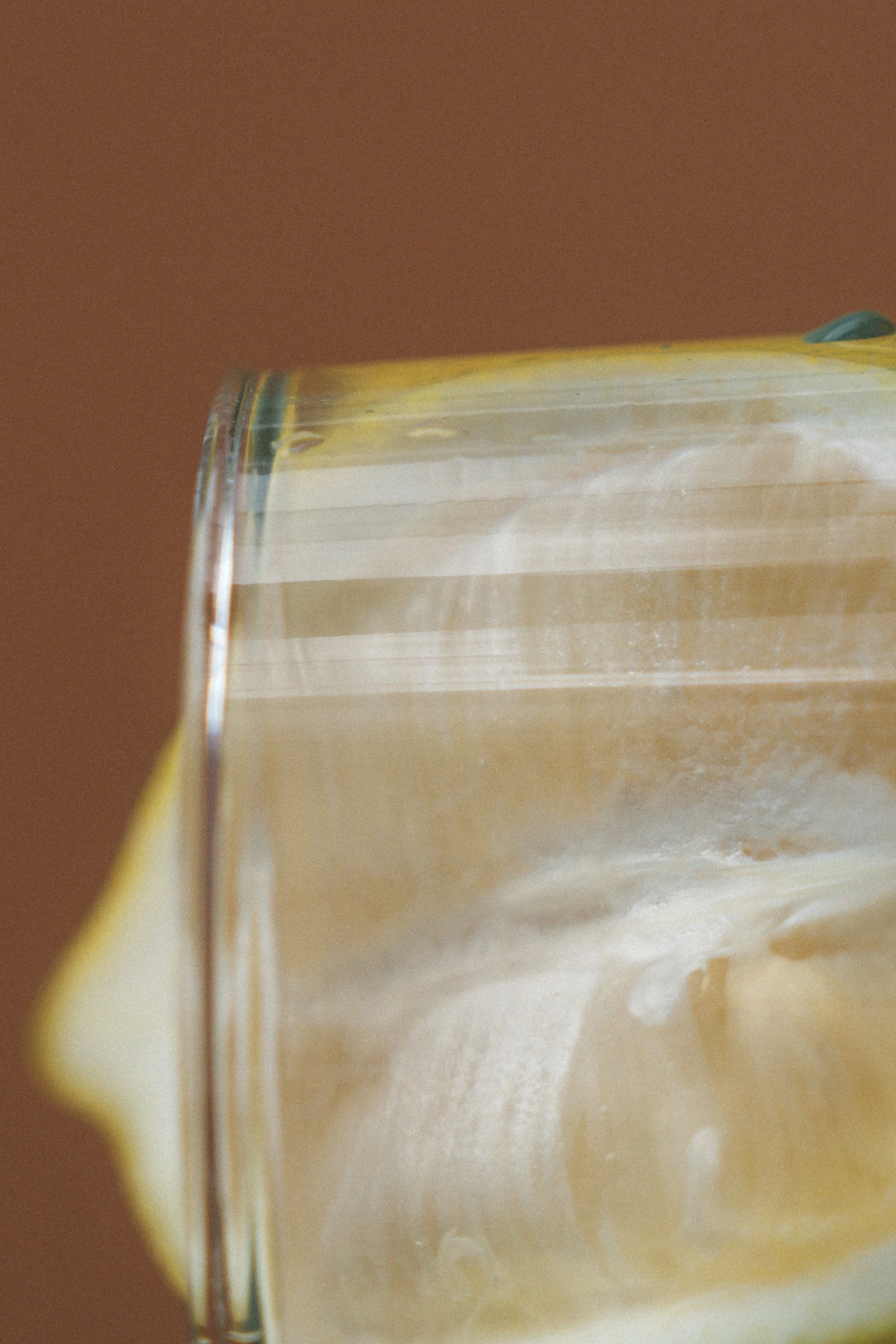 Close-up of a clear glass being filled with a light-colored, possibly creamy or milky liquid against a brown background.