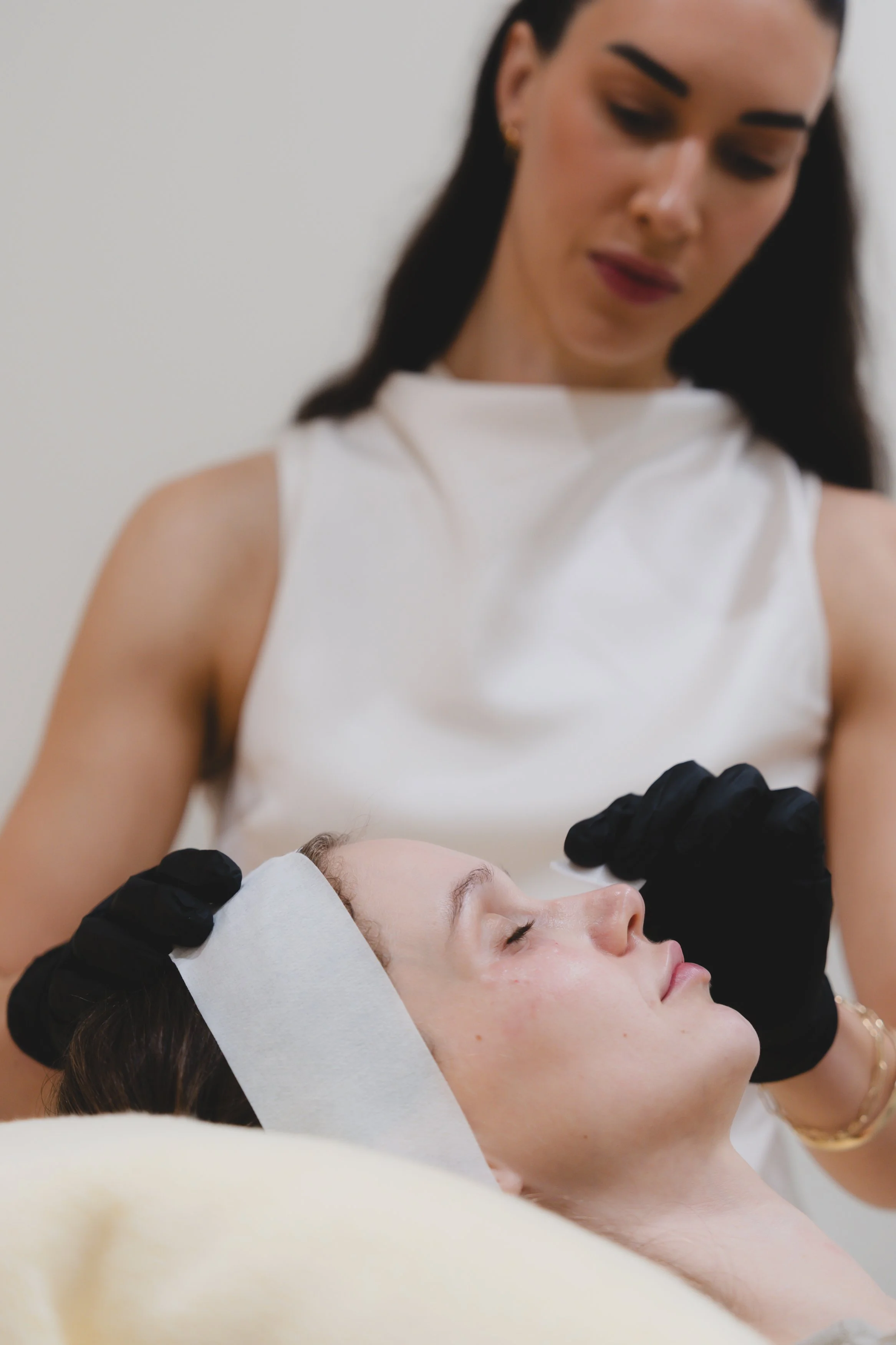 A woman lying on her back with closed eyes, receiving a facial treatment from a professional wearing black gloves, in a spa or clinic setting.
