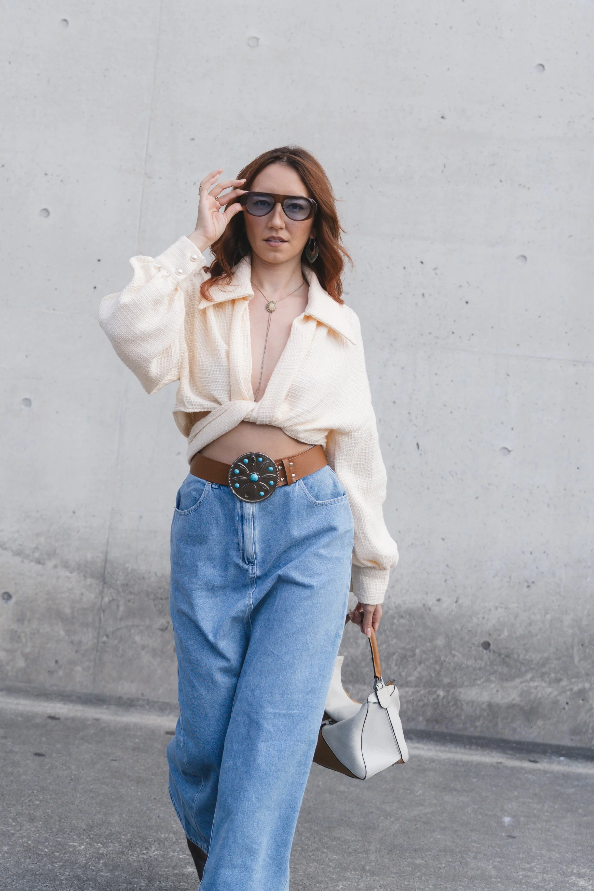 A woman with red hair wearing dark sunglasses, a cream-colored cropped shirt, and high-waisted blue jeans, holding a small white handbag while walking in front of a concrete wall.