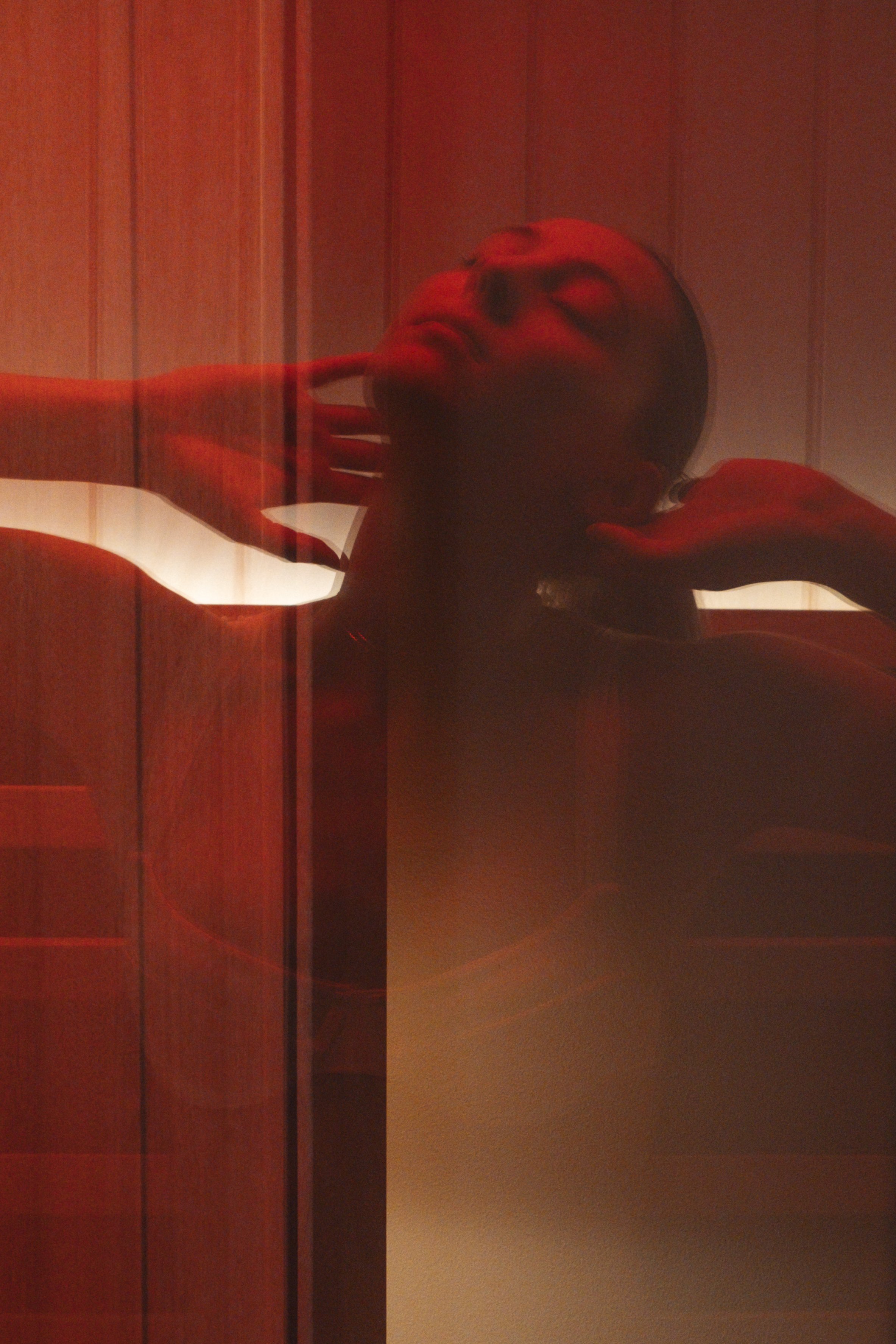 A woman with dark hair lying on a bed with her eyes closed, touching her neck, with vertical wooden wall panels in the background, illuminated by red lighting.