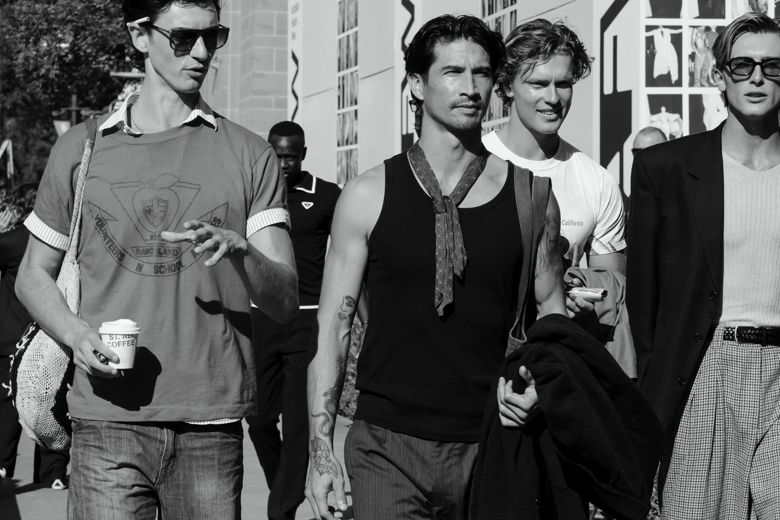 Black and white photo of a group of people walking outdoors. There are four individuals prominently visible, two women and two men, all dressed casually. The background includes other people and a decorated building.