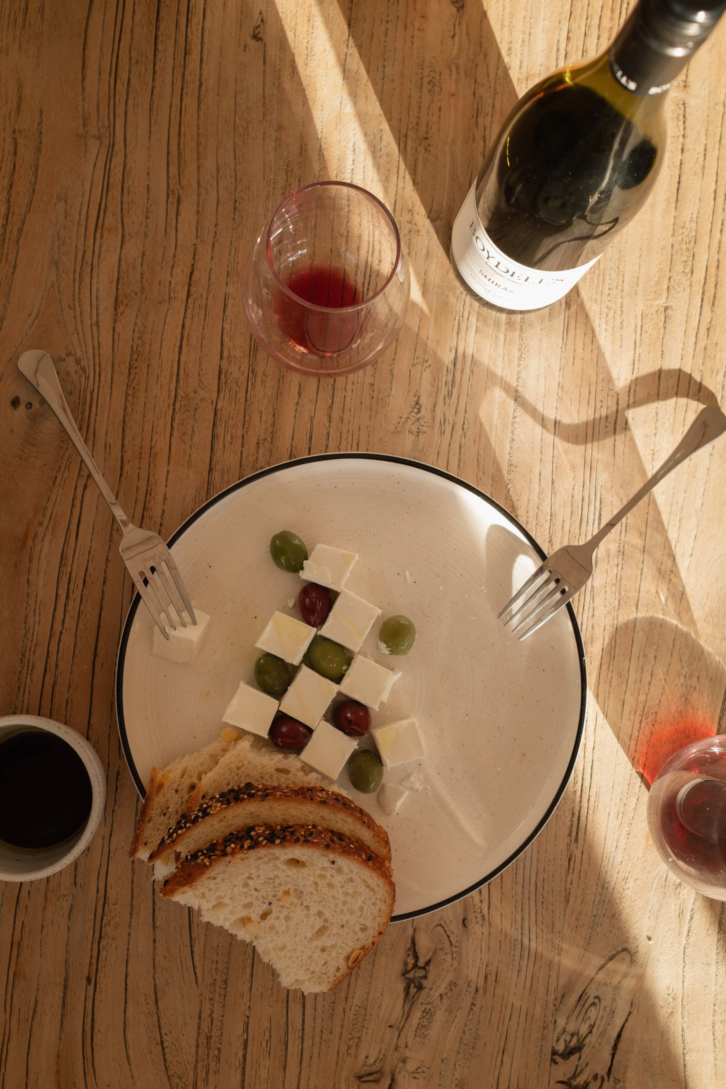 A plate of bread and cheese with grapes and olives, a glass of red wine, a bottle of wine, and some utensils on a wooden table.