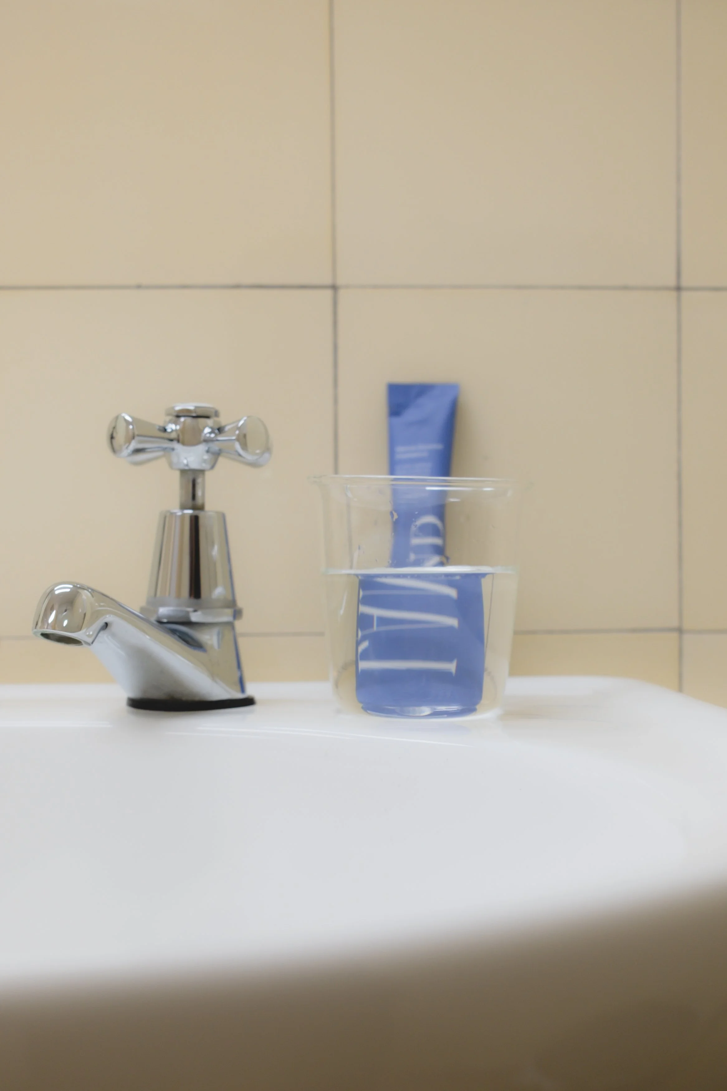Bathroom sink with a chrome faucet, a clear glass with water, and a tube of blue toothpaste in the background against tiled wall.
