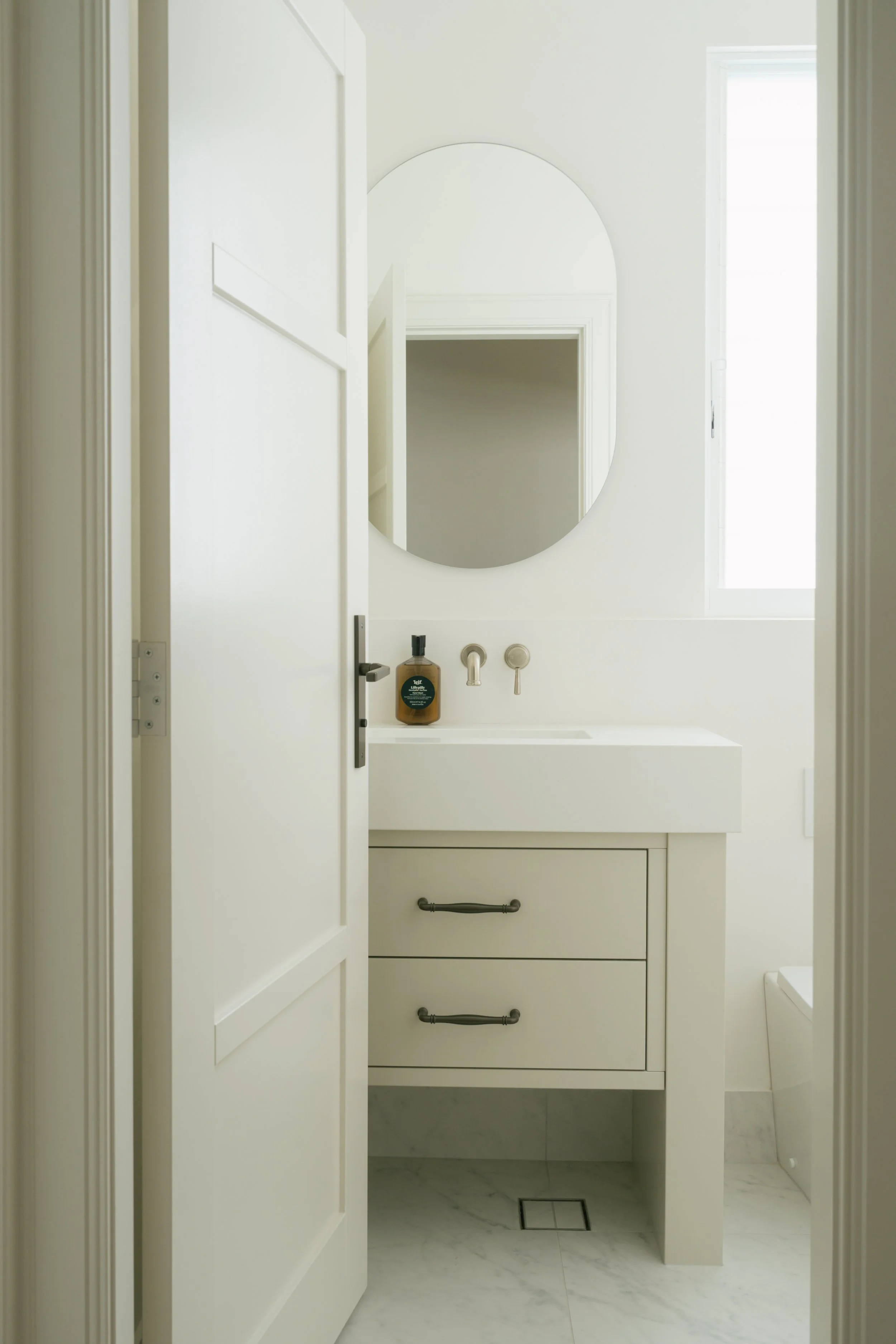 A minimalist bathroom with a white vanity, round mirror, and a bottle of hand soap on the sink, with natural light coming through a window.
