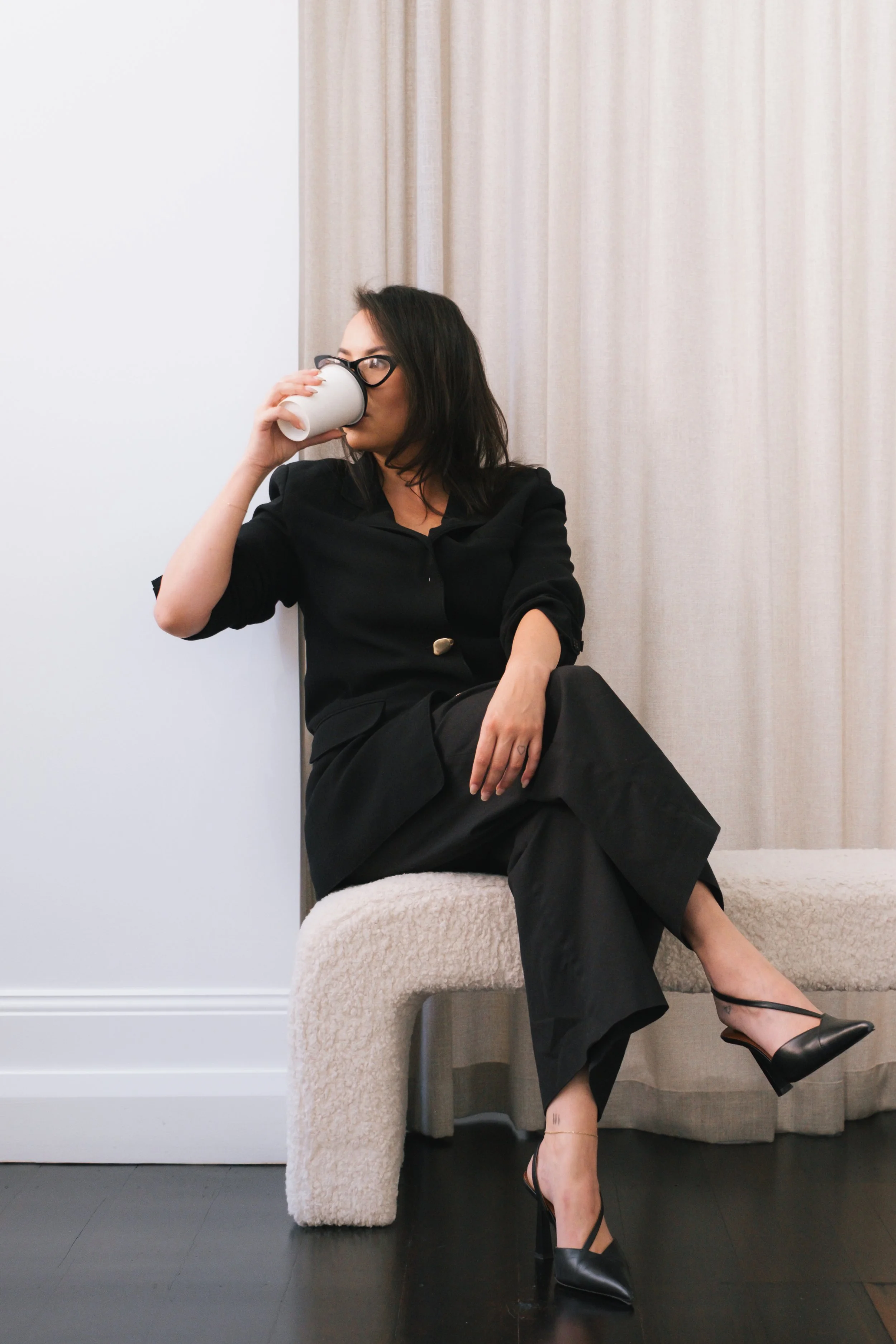 A woman with shoulder-length dark hair and glasses drinking from a white mug while sitting cross-legged and leaning against a beige curtain. She is wearing a black blazer and black pants, with black high-heeled shoes. She is seated on a white plush s