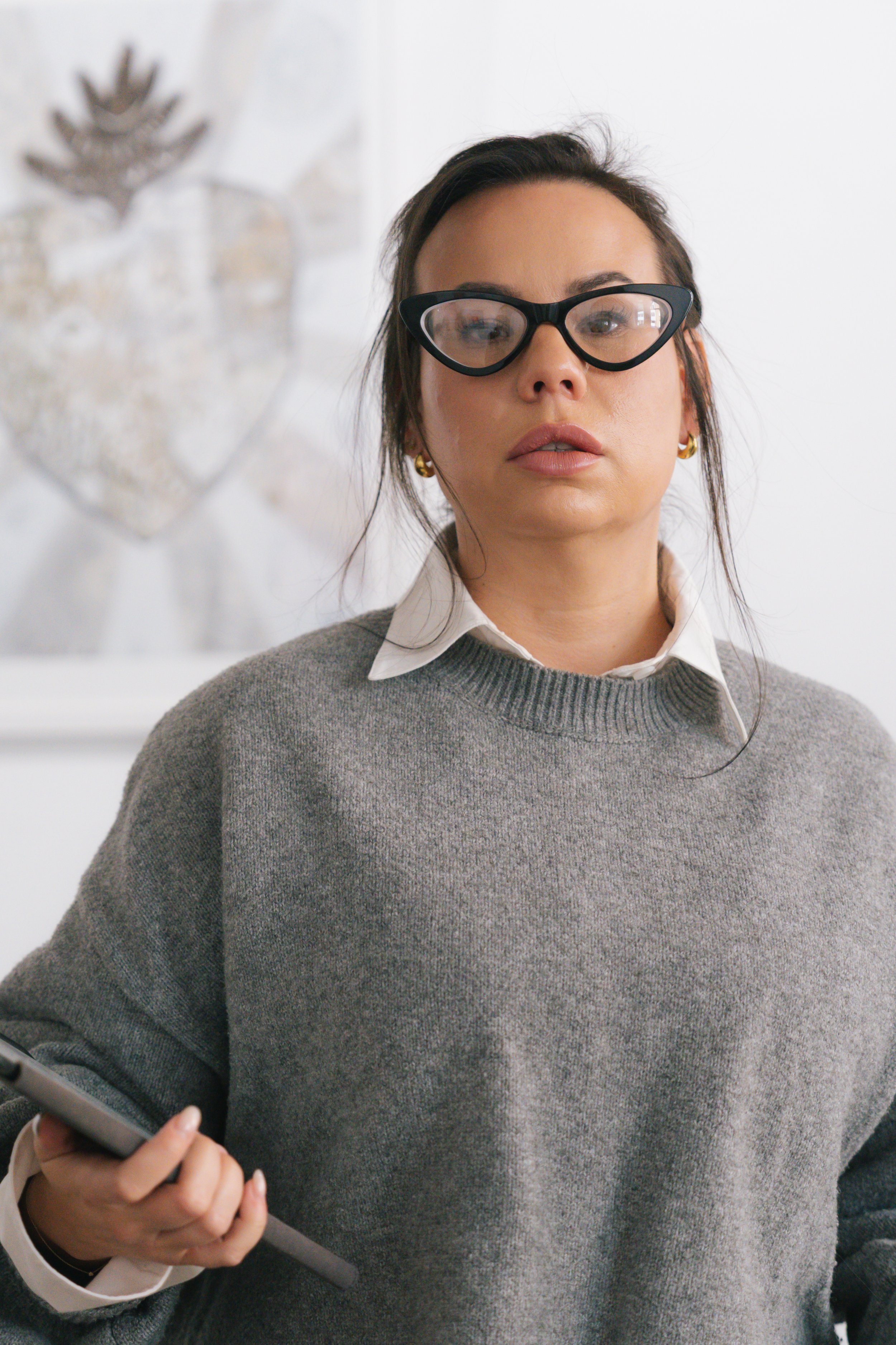 A woman with dark hair, wearing glasses, a gray sweater over a white collared shirt, and gold earrings, holding a smartphone in her hand.