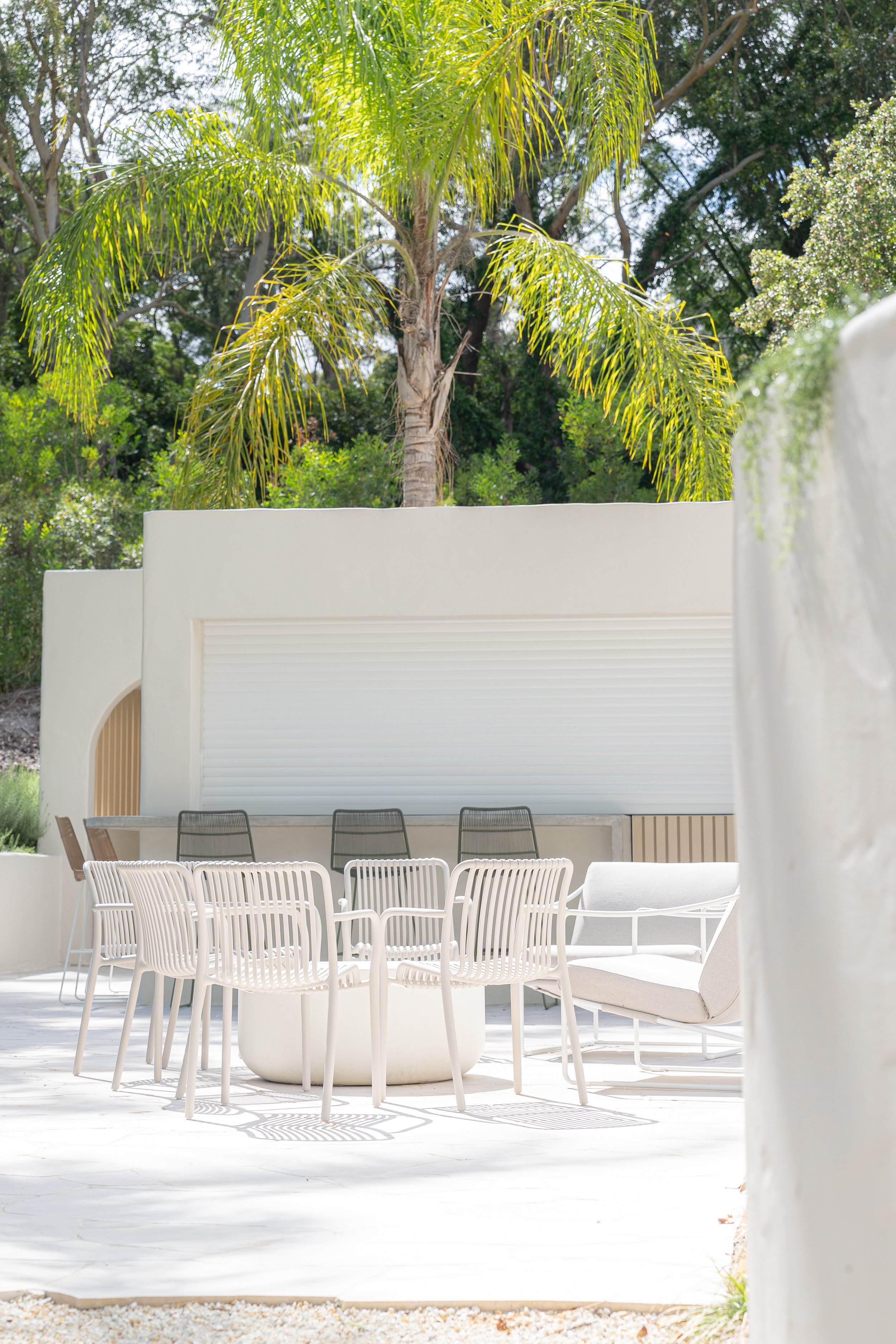 Outdoor patio with white modern dining chairs and a white curved sofa, surrounded by a white wall, with a background of green trees and a palm tree.