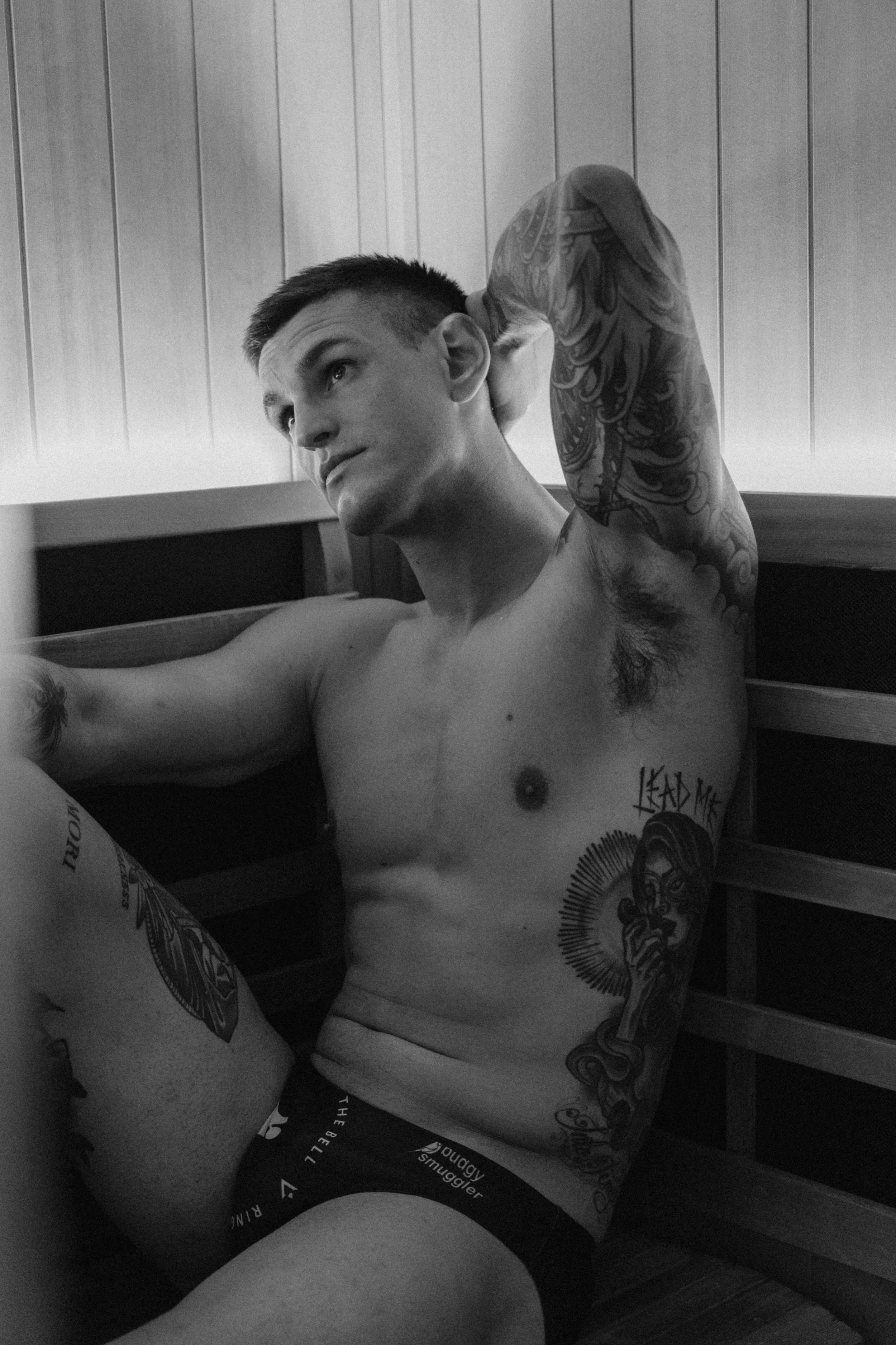 A shirtless man with tattoos on his arm and chest, sitting on the floor with a wooden wall behind him and a monitor in front, relaxing and looking away.