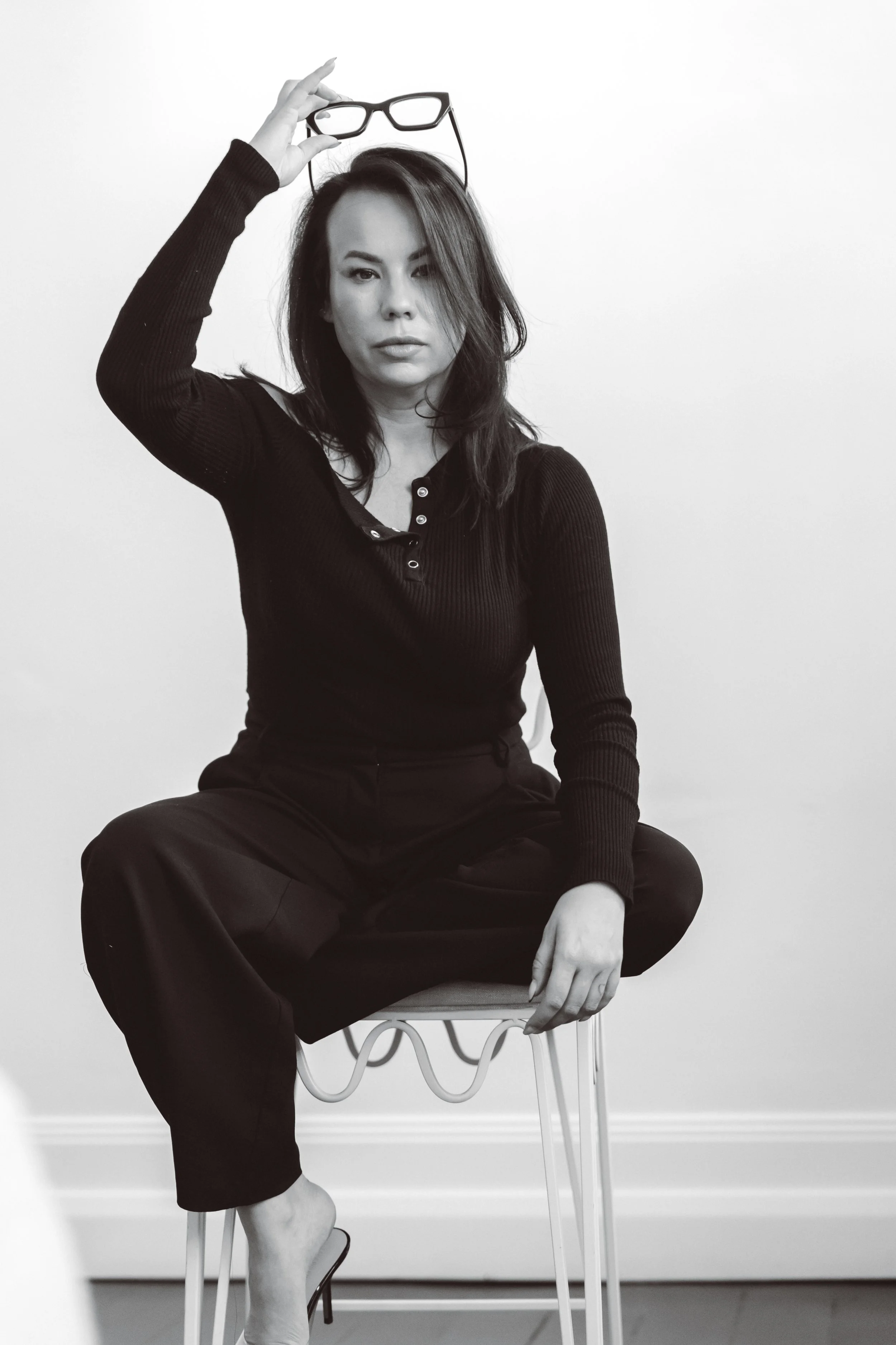 Black and white photo of a woman sitting on a chair, holding glasses in her right hand on top of her head, wearing a long sleeve black top and black pants, with one leg crossed over the other, in front of a plain white wall.