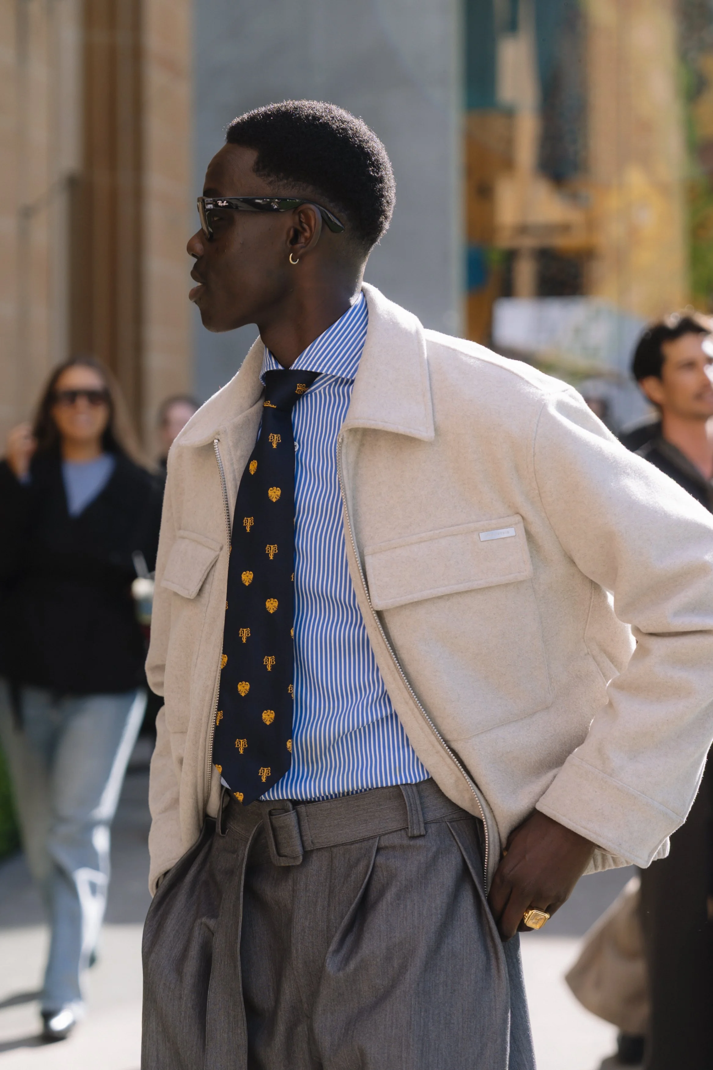A young black man wearing sunglasses, a blue and white striped shirt, a navy tie with gold pattern, a light-colored jacket, and gray pleated trousers. He is in an outdoor setting with other people in the background.