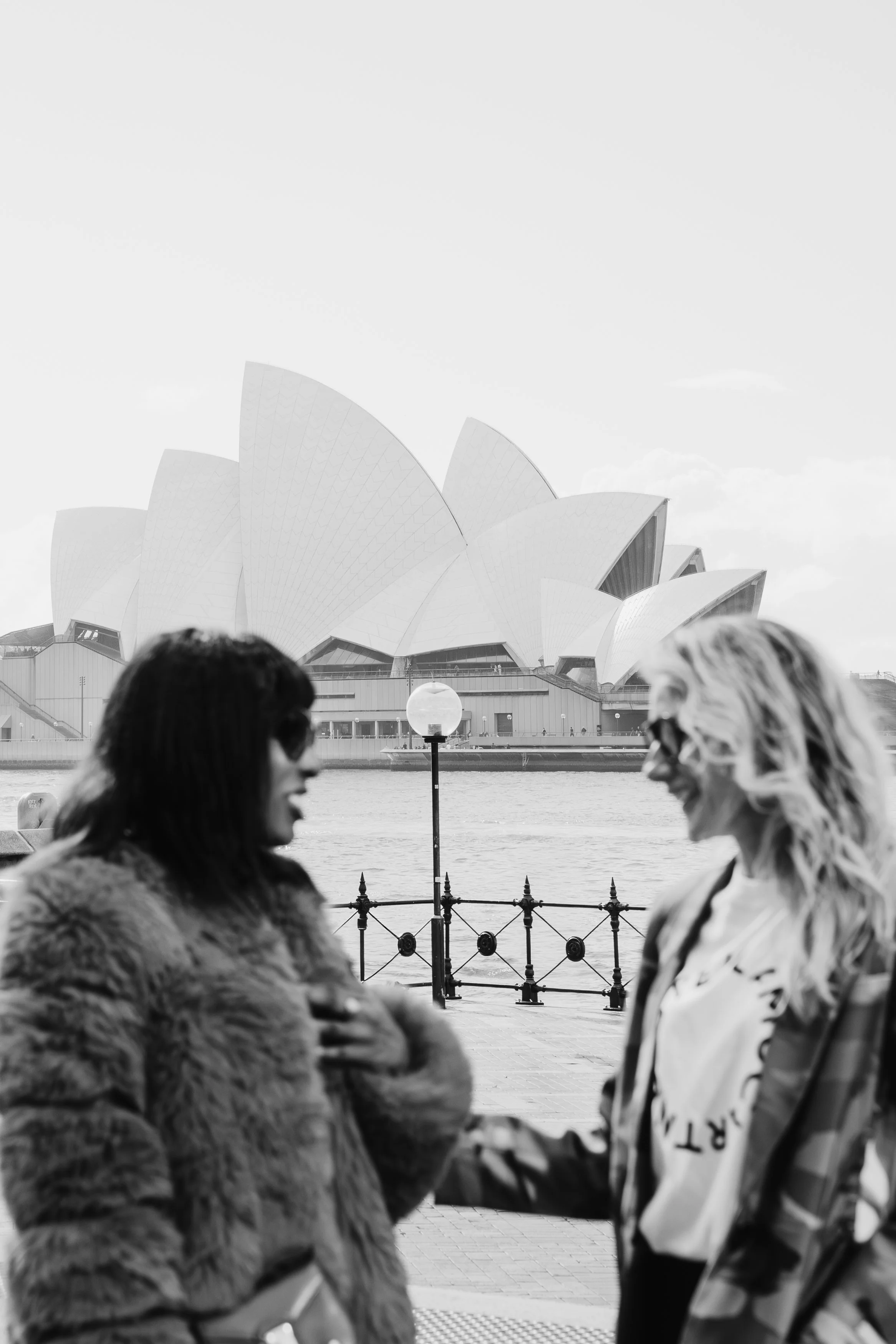 Two women standing and talking near a railing with the Sydney Opera House in the background, in black and white.