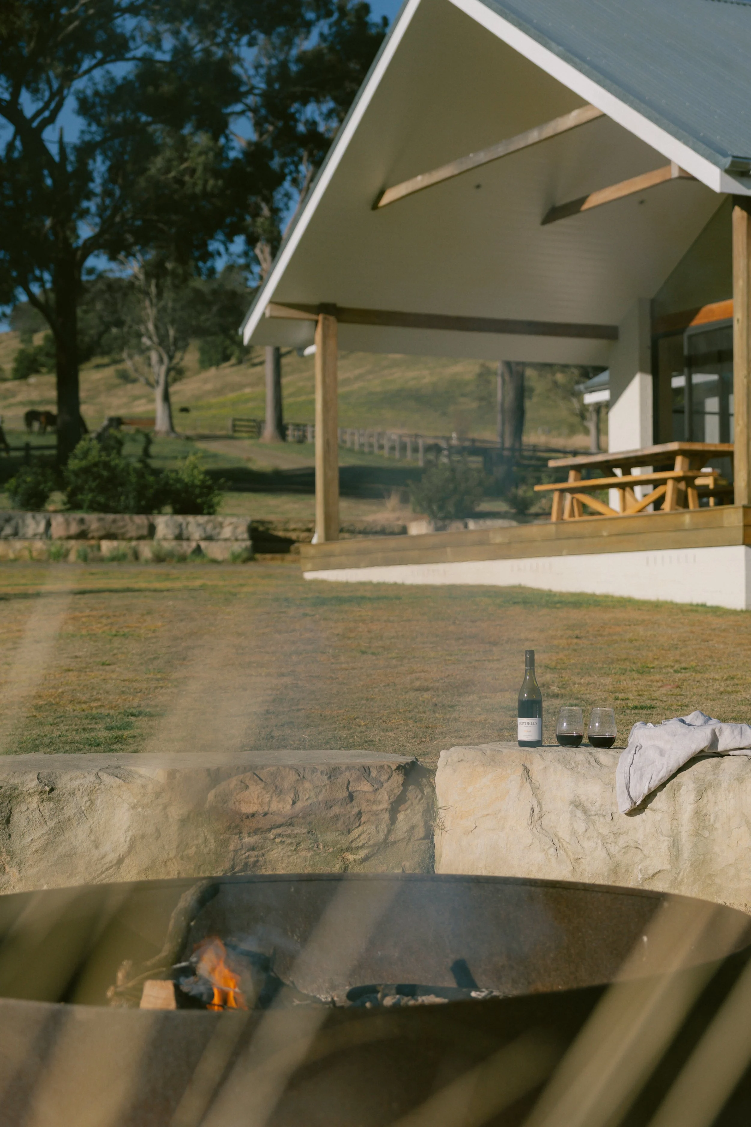 A bottle of red wine, two wine glasses filled with red wine, and a white cloth on a stone surface in an outdoor setting with a house, picnic table, trees, and a field in the background, viewed from inside a vehicle near a fire pit.