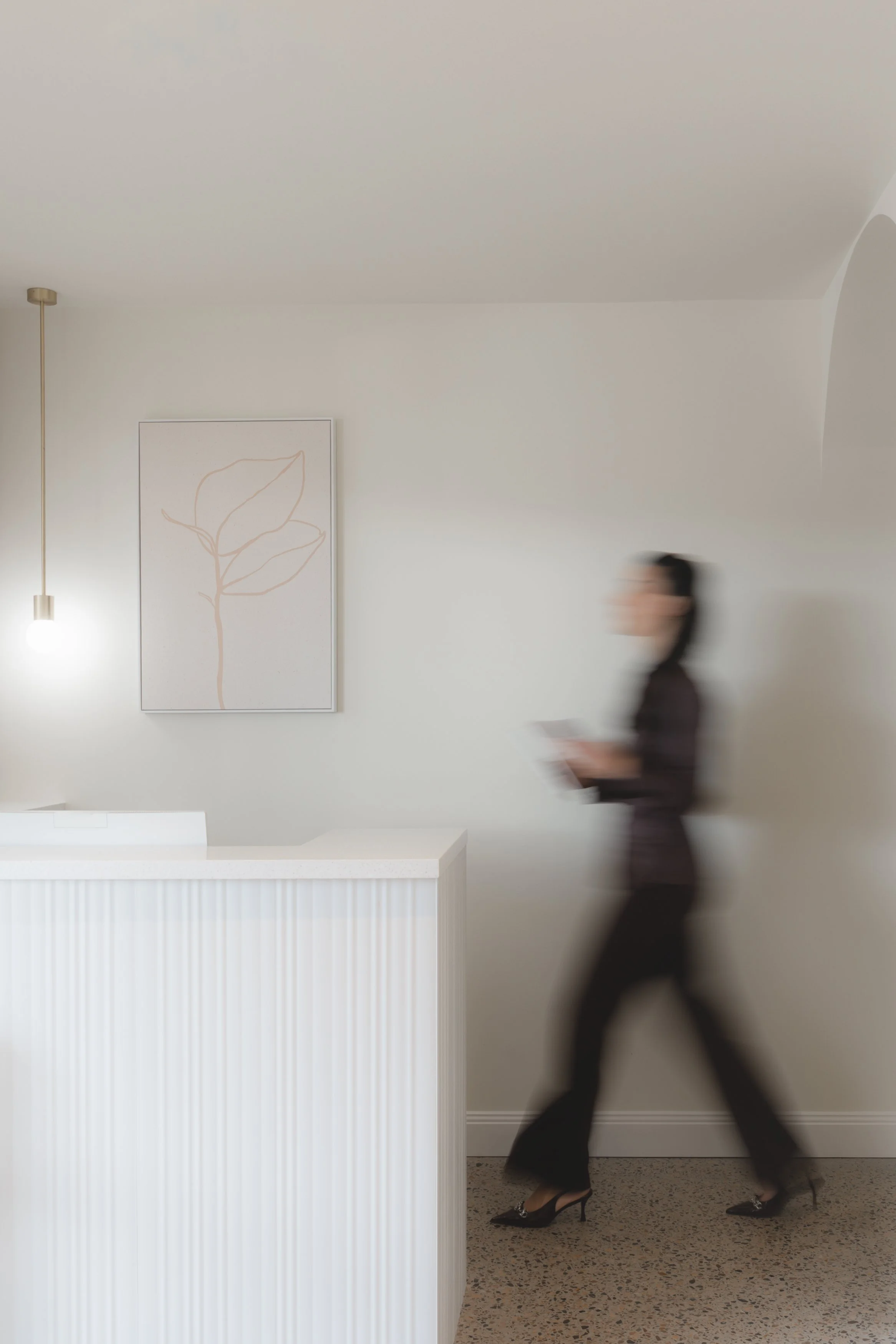 A woman in black attire and high heels walking past a white panel with a linear art print of a leaf on a plain white wall, with a modern hanging light and terrazzo flooring.