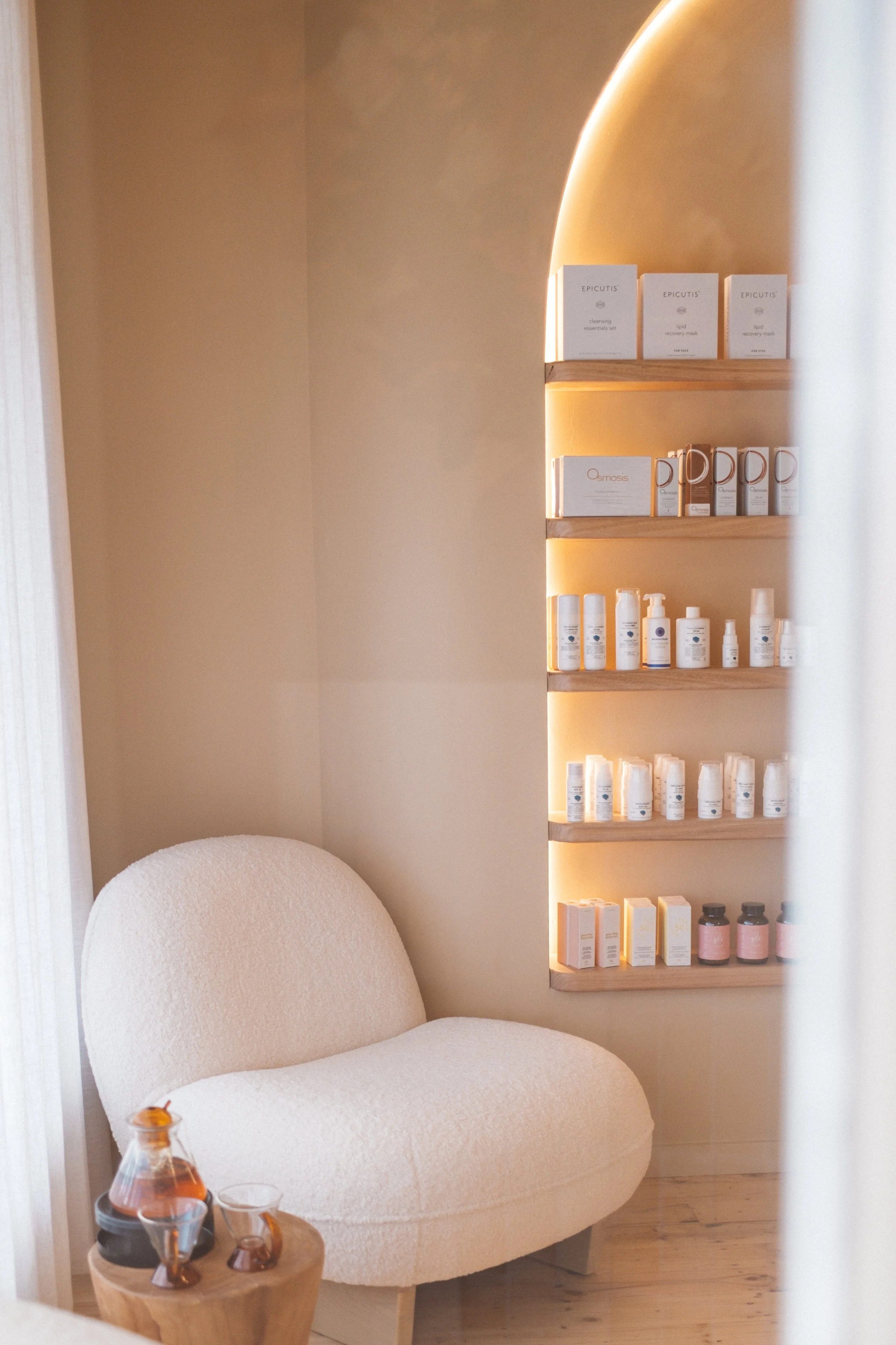 A cozy corner of a spa or wellness center featuring a white, plush, curved armchair next to a small wooden side table with bottles and glasses. Behind, there's a wooden shelf built into a wall with soft, warm lighting, displaying various skincare pro