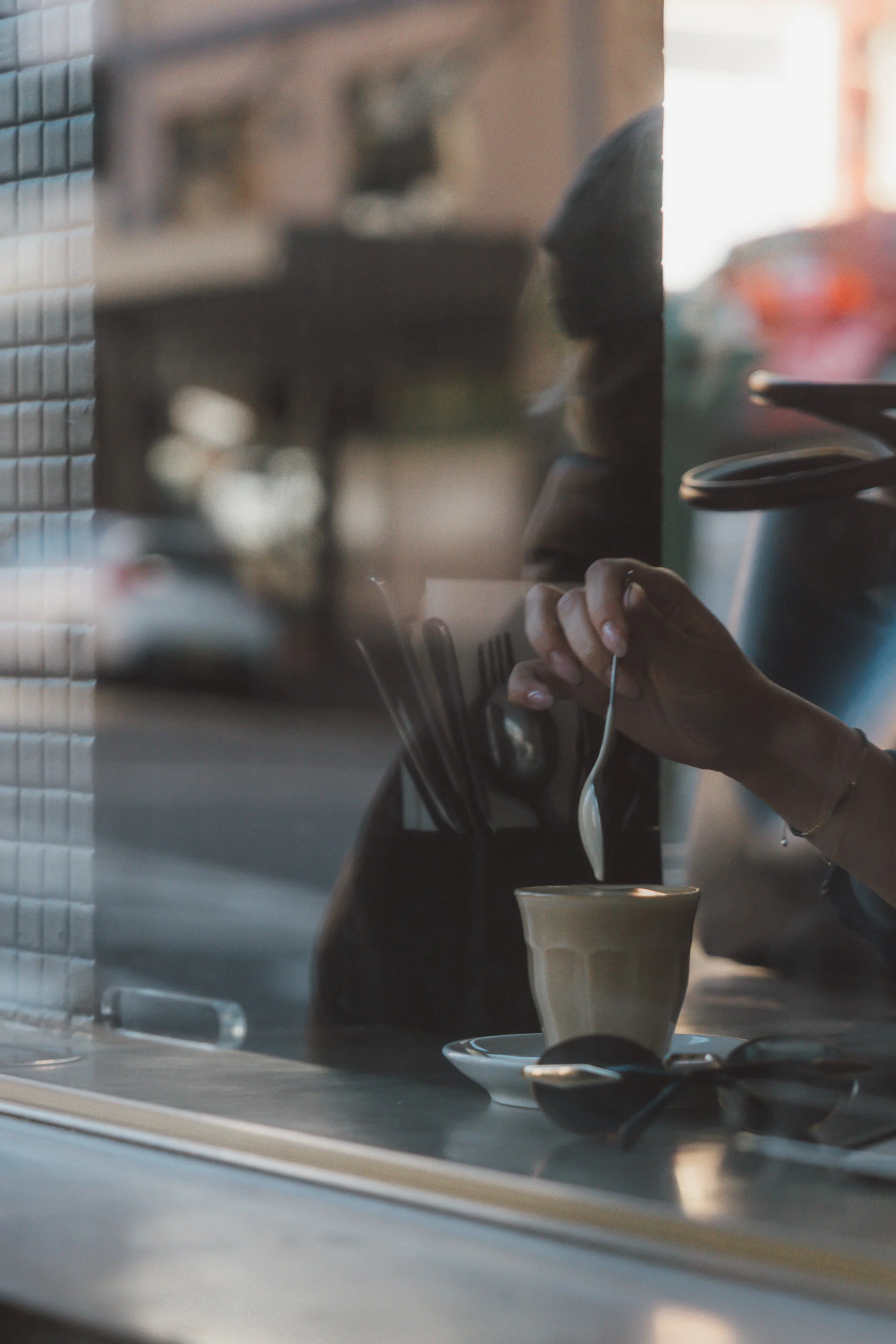 A person wearing glasses is pouring a frothy beverage into a glass on a table in a cafe or restaurant, with a reflection visible on a glass window in the foreground.