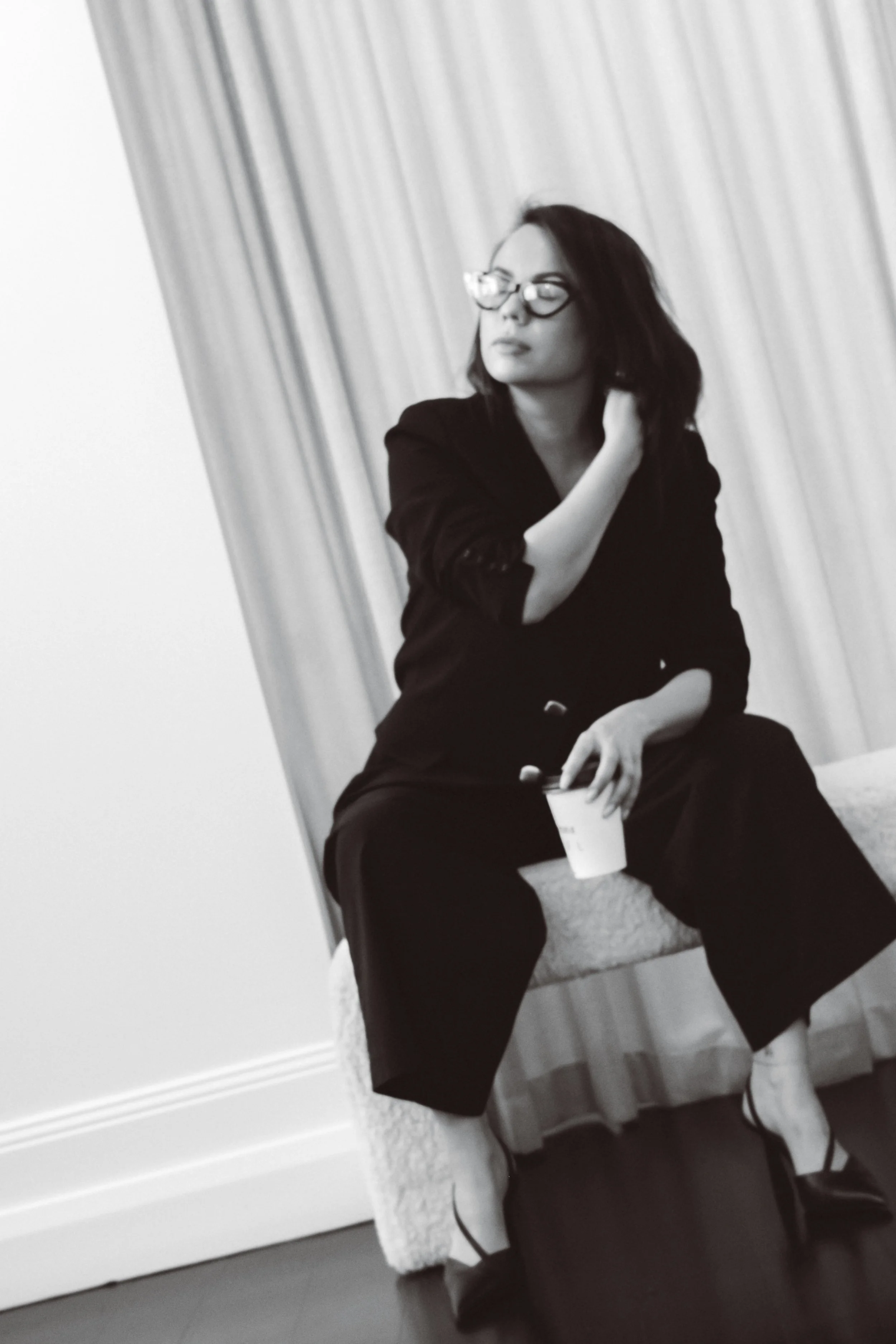 A woman with shoulder-length dark hair and glasses, dressed in a black blazer, sitting on a stool with a cup in her hand, in front of a curtain background.
