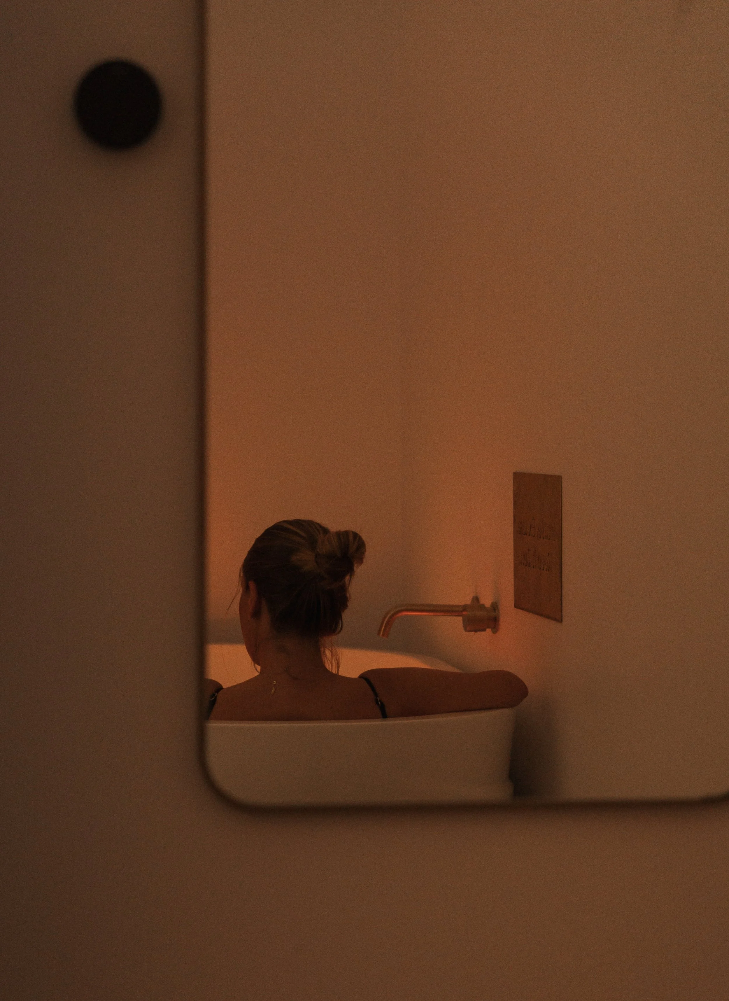 A woman with brown hair styled in an updo sitting in a bathtub, viewed through a mirror, with a wall-mounted faucet and a wooden sign on the wall.