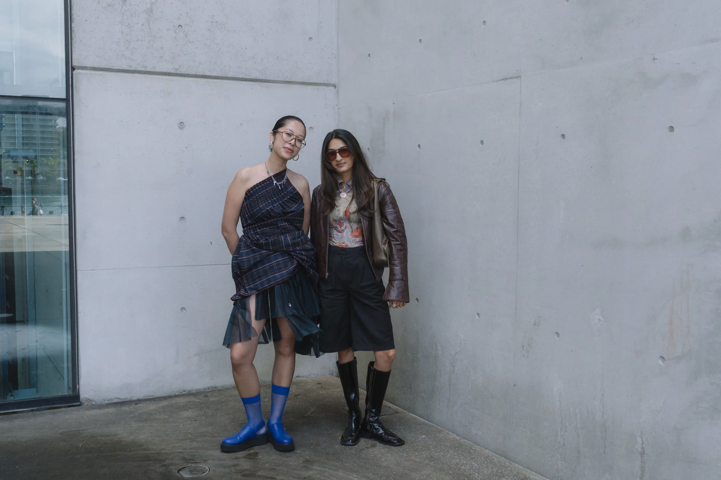 Two women standing close together against a concrete wall outside. The woman on the left has glasses, her hair in a bun, and is wearing a strapless plaid dress with sheer black overlay and blue socks with black shoes. The woman on the right has long dark hair, sunglasses, a patterned shirt with a brown leather jacket, and knee-high black boots.