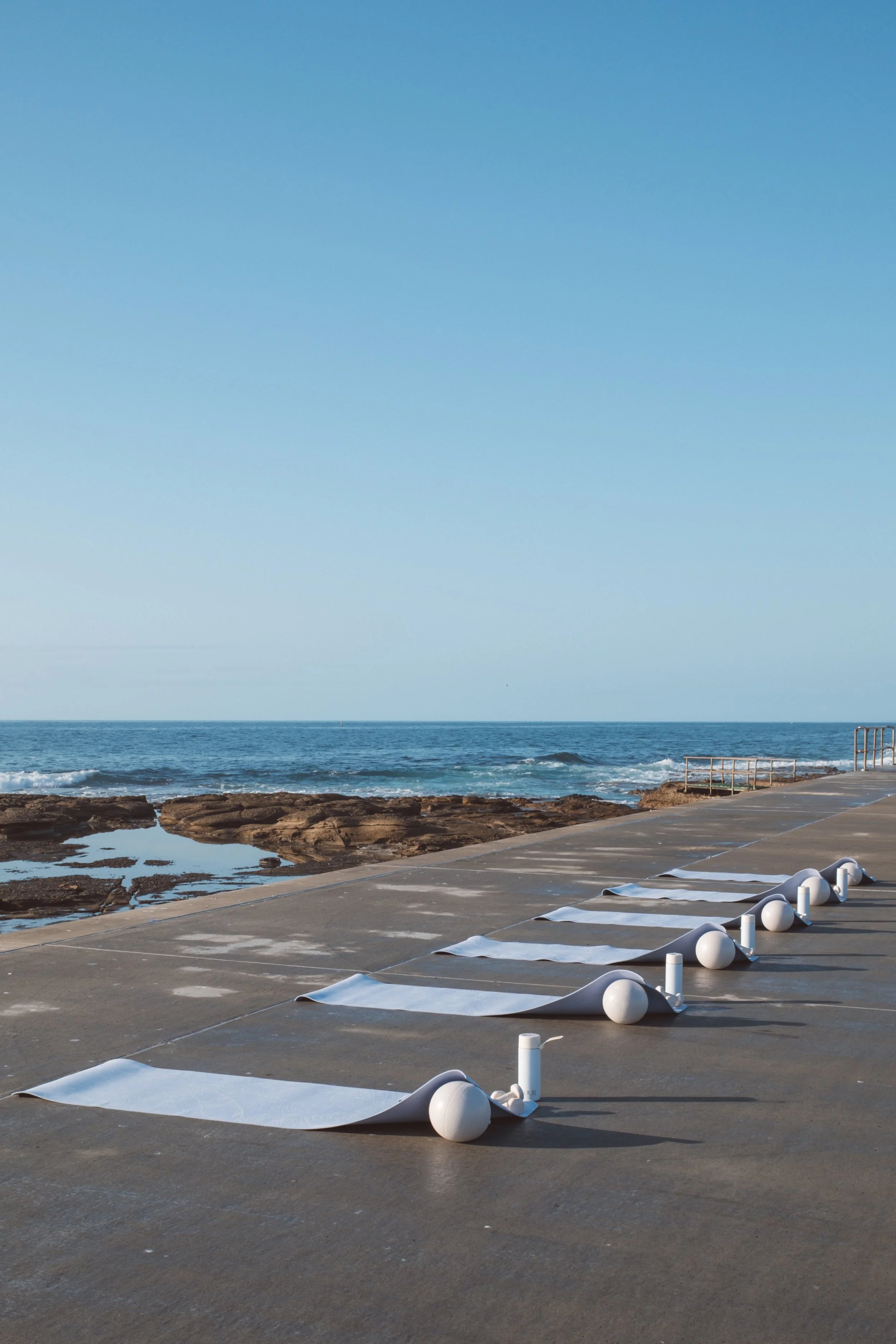 Empty outdoor stage with white paper and balloons on the ground near the shore, calm ocean waves, rocks, blue sky.