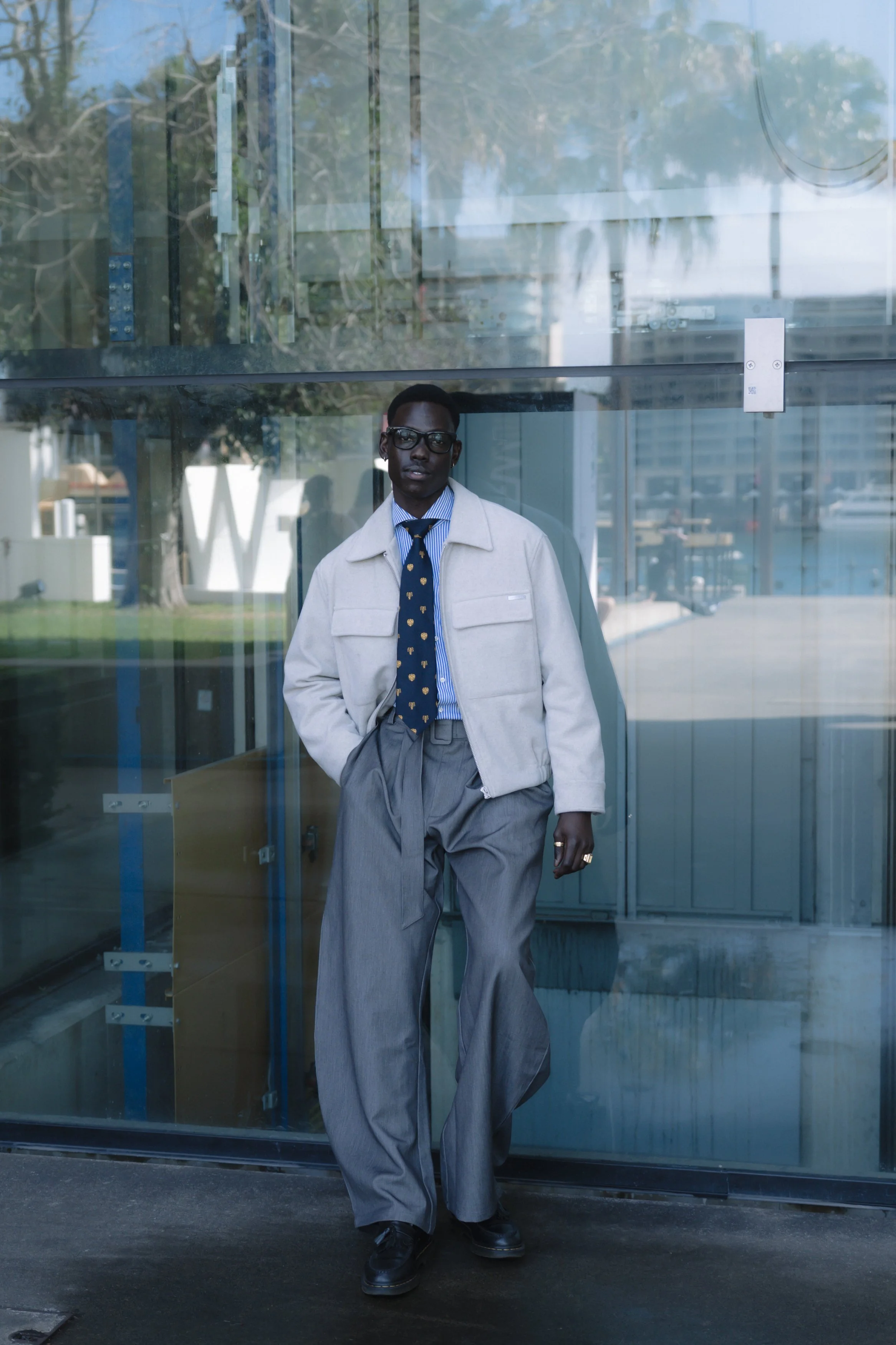 A man wearing glasses, a white jacket, a blue striped shirt, and a navy tie with yellow motifs, standing in front of a glass building.