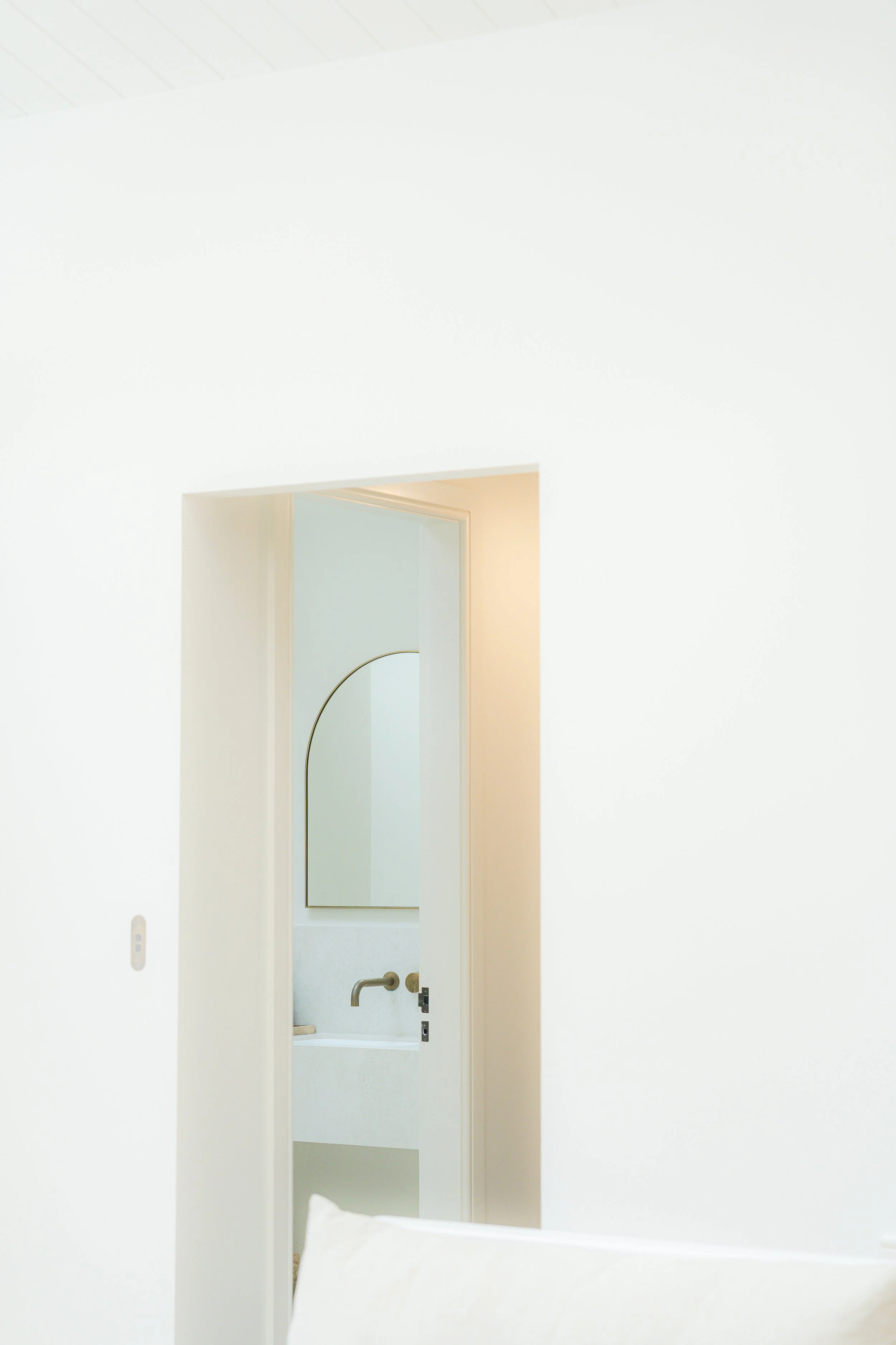 White-walled bathroom with a mirror, sink, and faucet, partially visible through a doorway.