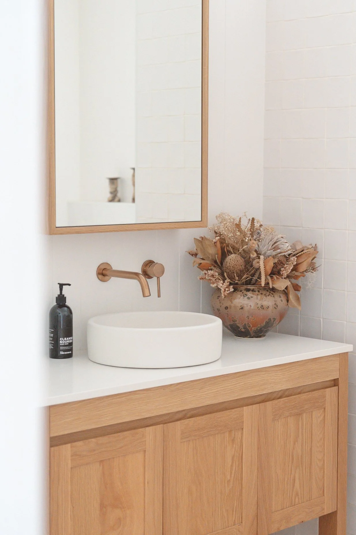 A bathroom vanity with a white vessel sink, a black soap dispenser, a gold wall-mounted faucet, a large ceramic vase with dried flowers on a white countertop, a wooden cabinet, a mirror with a wooden frame, and white tiled walls.